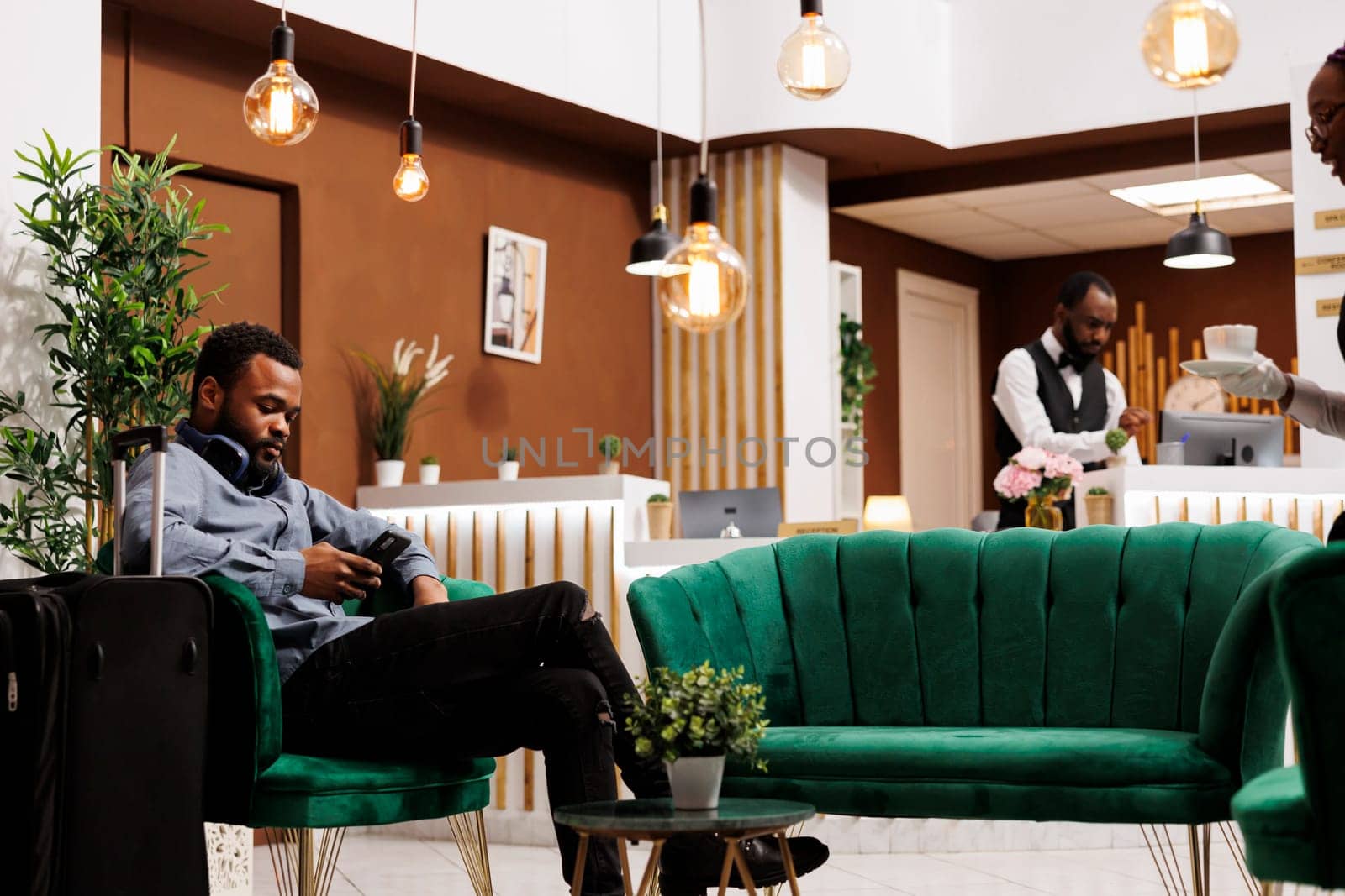 Black guy traveler sitting in hotel lobby with smartphone making mobile check-in. Young African American man tourist with luggage using phone texting sms message, waiting for room