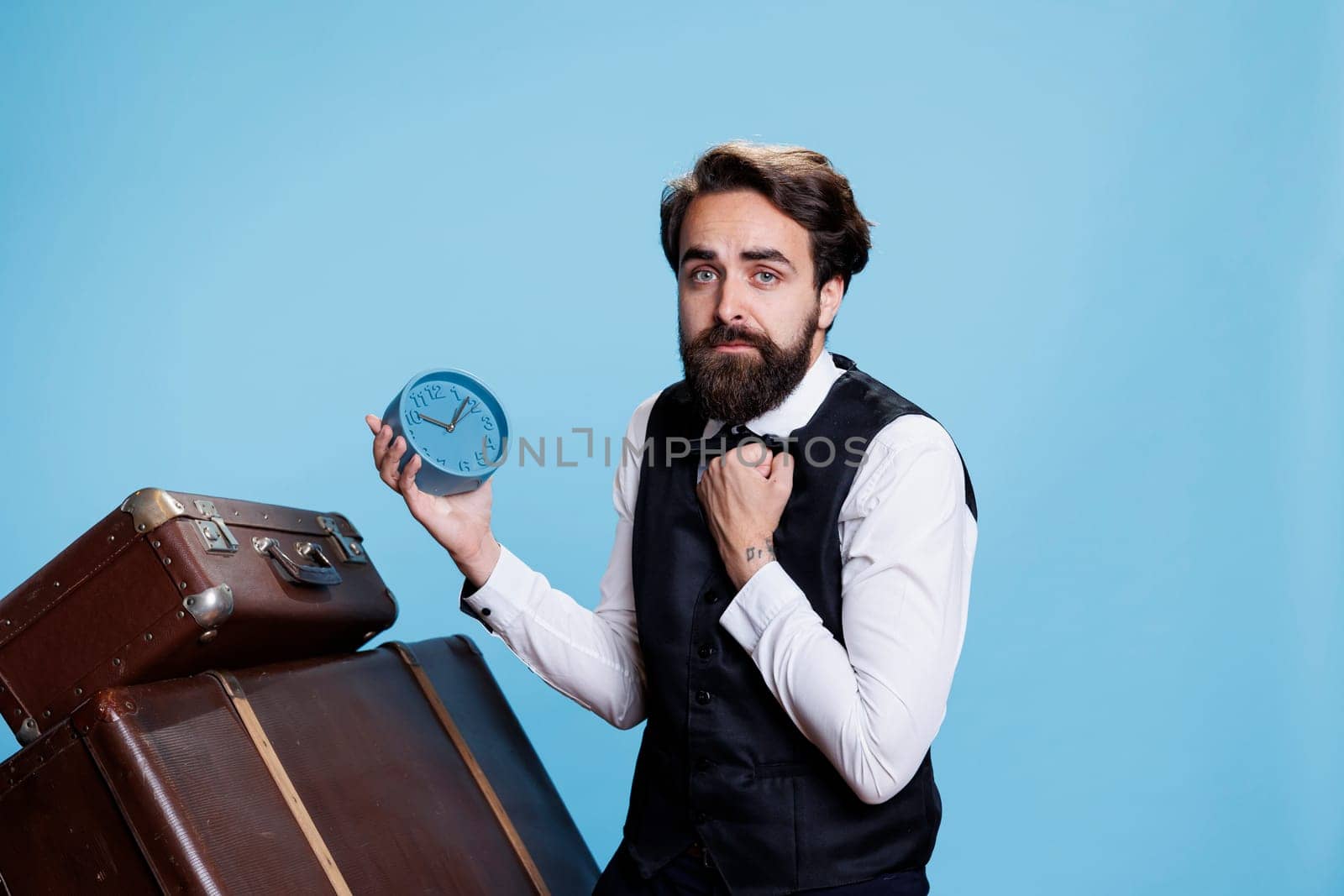 Classy bellboy uses clock to see time and posing next to trolley bags, professional person hates to be late. Employee in travel industry looking at watch to tell hour before hotel guests arrive.