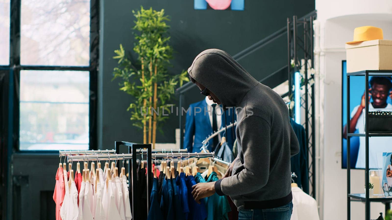 African american thief robbing clothing store, analyzing racks with stylish clothes. Robber trying to steal fashionable merchandise, wearing sunglasses and hood not to be recognized