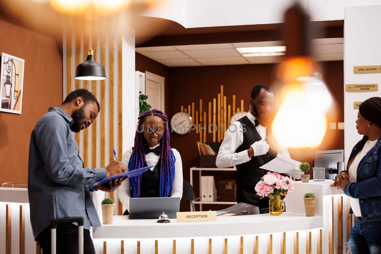Young African American guy hotel guest filling up registration form at front desk. Female receptionist checking in guests, collecting personal information from tourists during check-in procedure