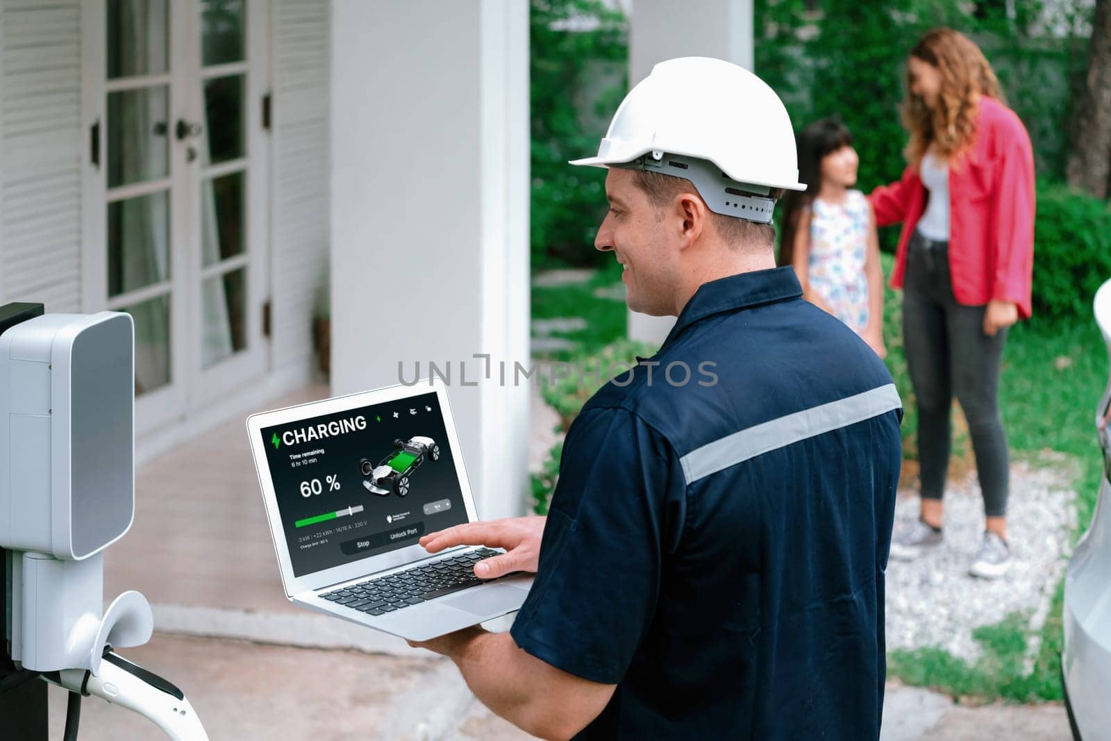 Qualified technician working on home EV charging station installation, making troubleshooting and configuration setup on charging system with laptop for EV at home with the family present. Synchronos