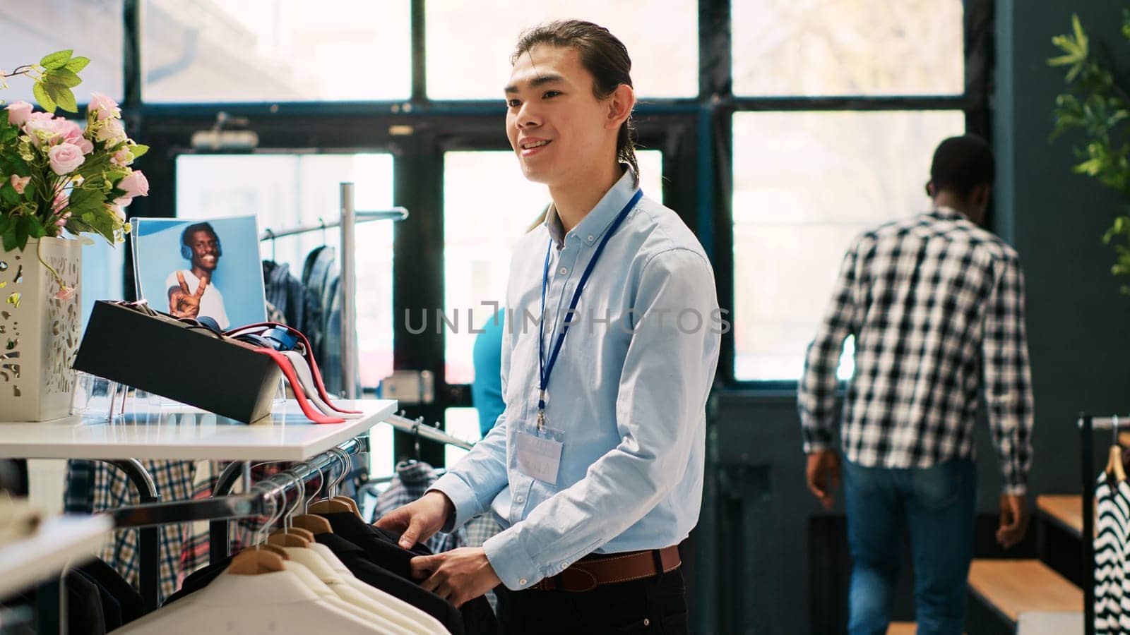 Asian manager preparing store for opening, arranging stylish clothes on hangers in shopping centre. Employee looking at fashionable merchandise, checking price in modern boutique