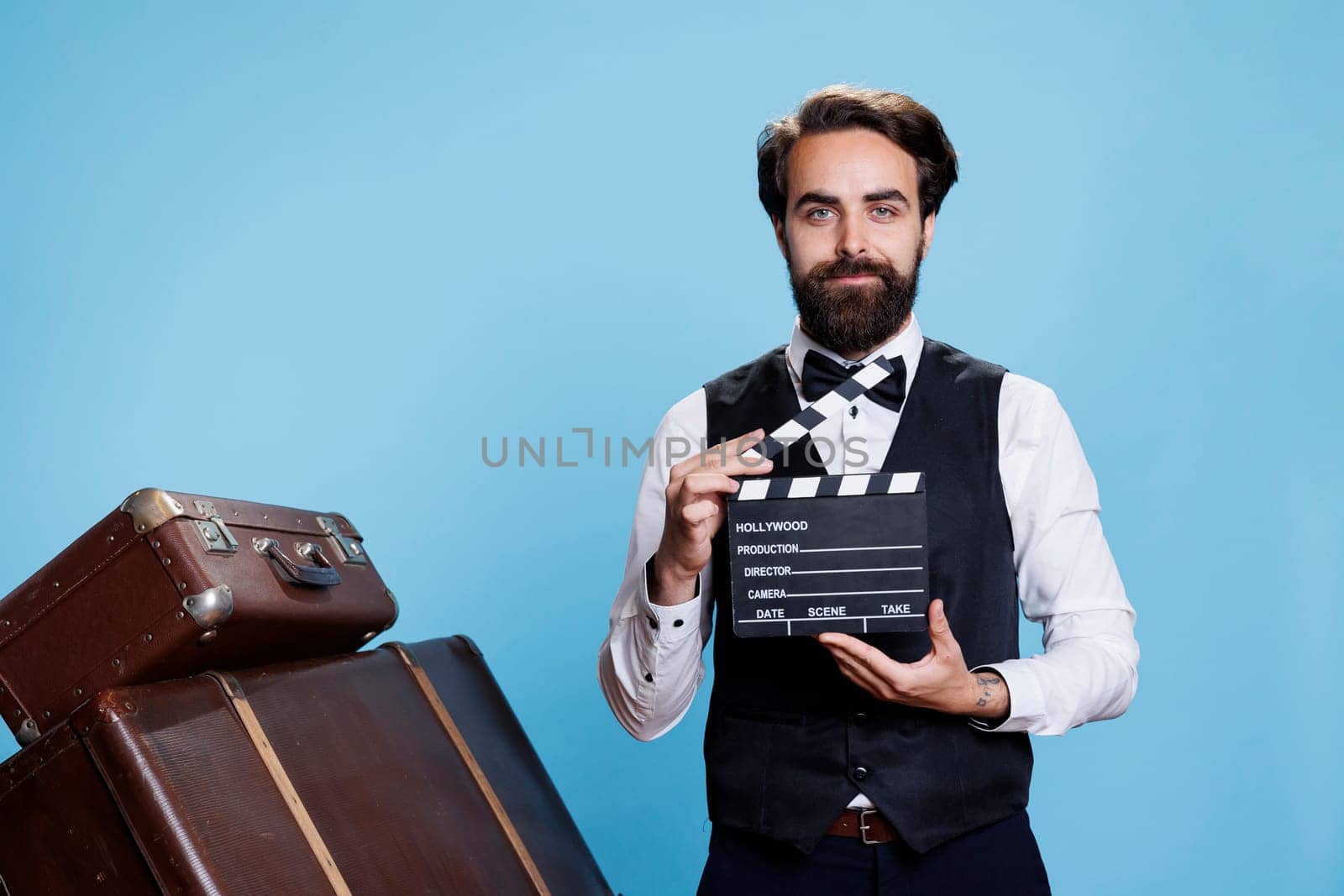 Doorkeeper clapping filmslate on camera, saying action to cut scene for fun against blue background. Classy hotel bellboy pretending to work in movie production using clapperboard.