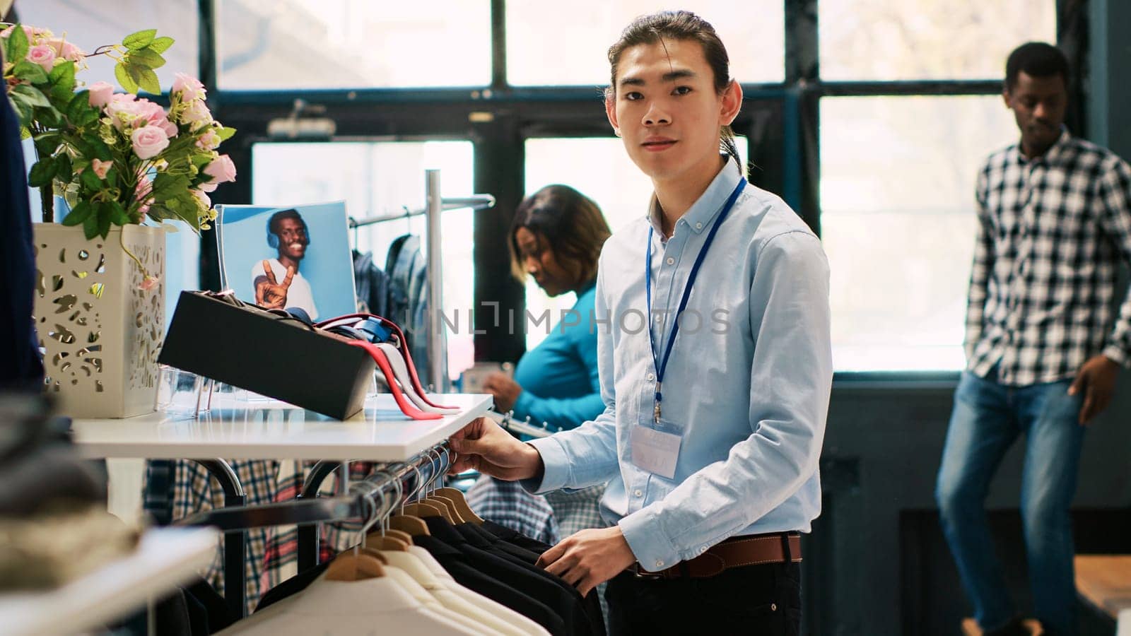 Asian worker arranging new fashion collection, putting hangers with fashionable merchandise on racks in modern boutique. Employee preparing store for opening, working with stylish clothes