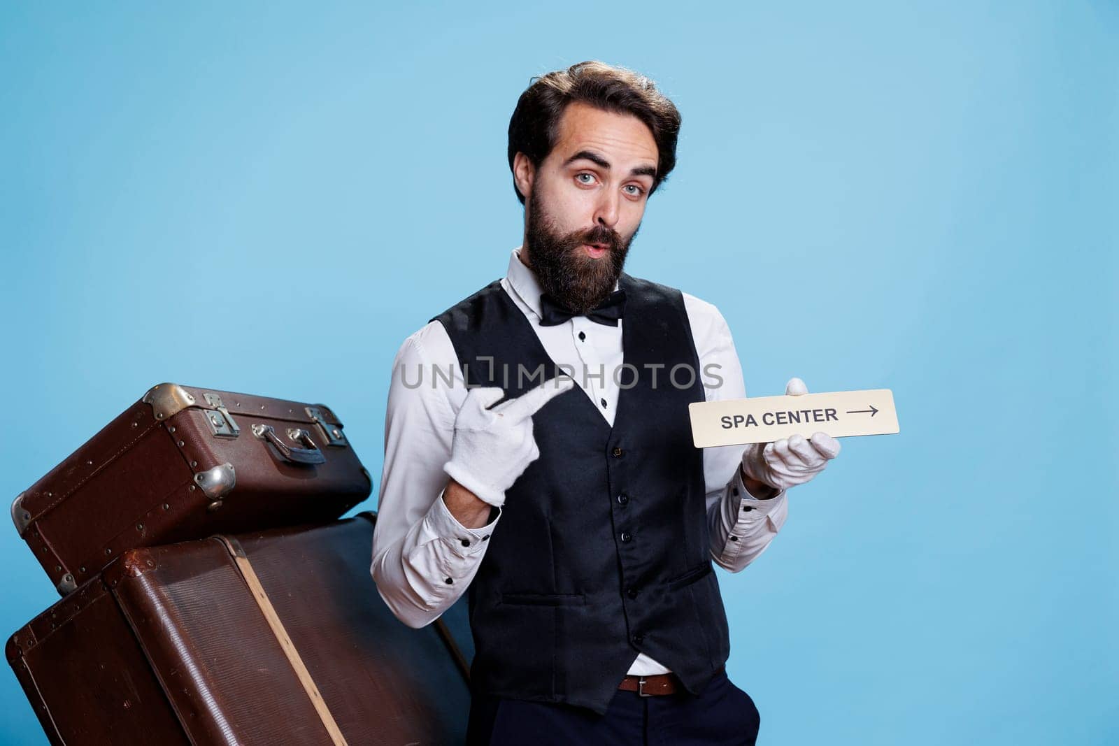 Confident bellboy uses spa center sign to direct guests and show all hotel locations, hospitality industry. Stylish skilled worker holding indicator or pointer for the relaxation area.