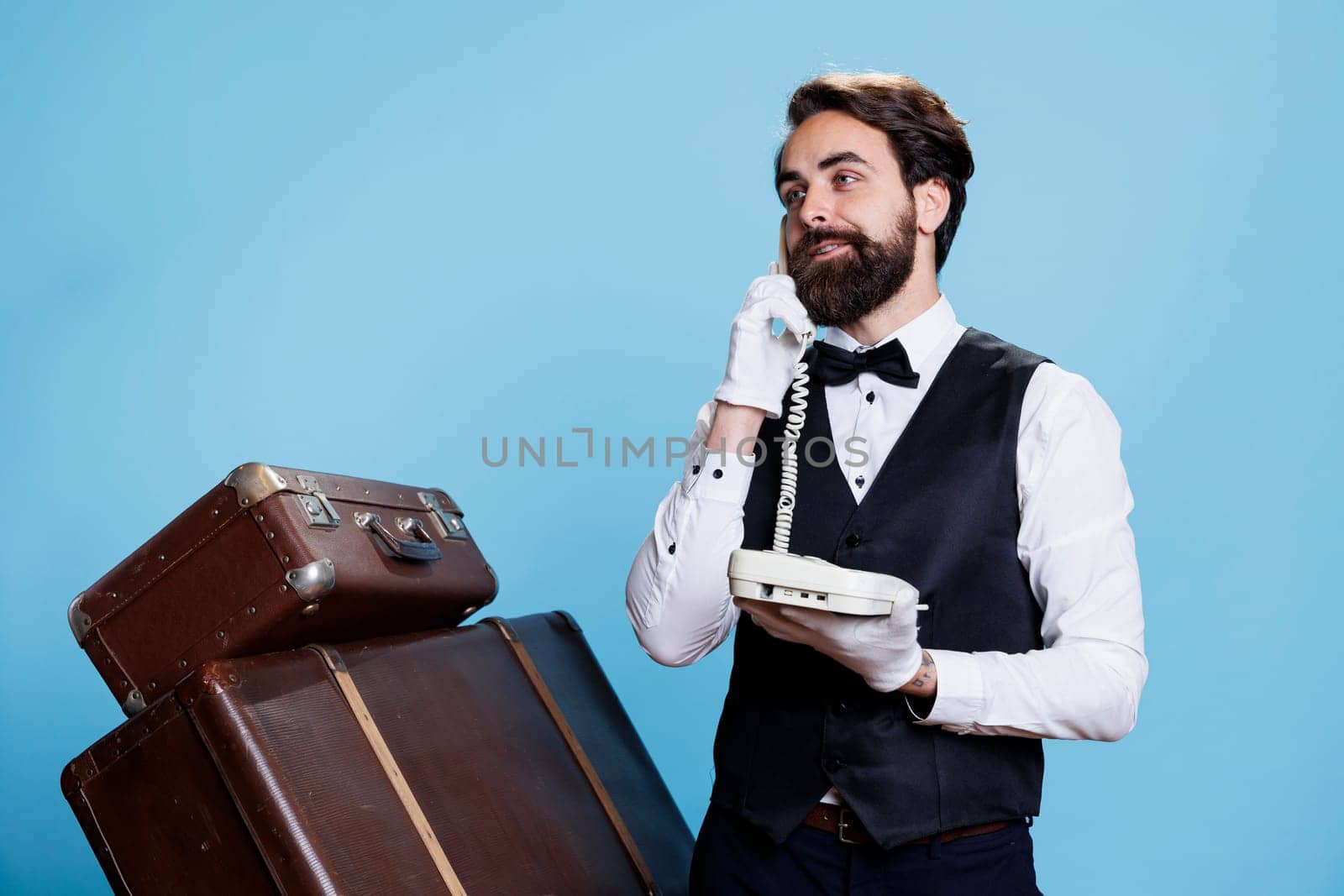 Stylish bellboy answers landline phone in studio, talking to hotel guests about accommodation. Skilled confident doorman using telephone with cord to take calls, vintage luxurious job.