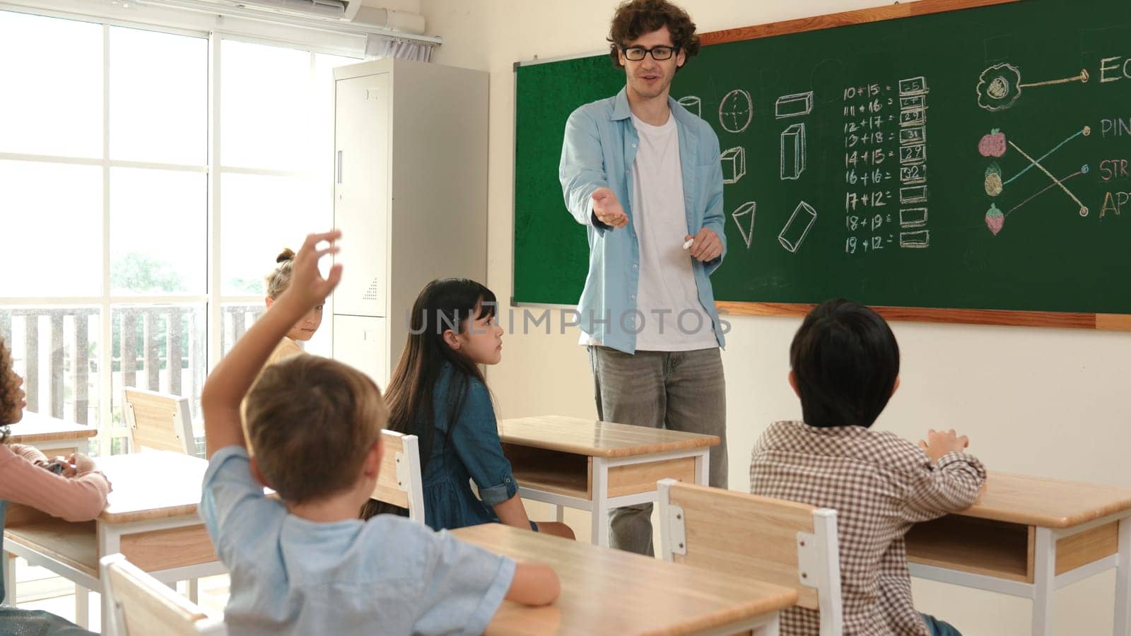 Energetic asian boy raised his hand and volunteer to write answer on blackboard. Teacher ask for volunteering to answer question while happy student receive chalk and walk in front of class. Pedagogy.