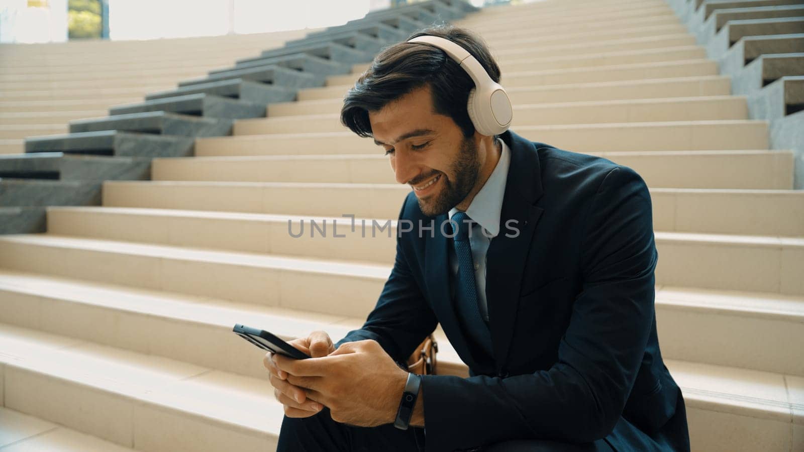 Smiling business man listening music while looking at mobile phone. Project manager holding phone while enjoy hip-hop song at stairs. Caucasian leader listen relax sound while plan project. Exultant.