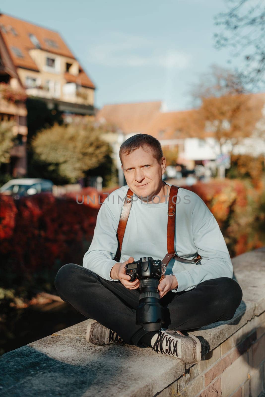 Man Sitting in Old European City And Holding Photo Camera. Contemporary Stylish Blogger And Photographer. Handsome man taking a selfie on a trip in Europe. by Andrii_Ko