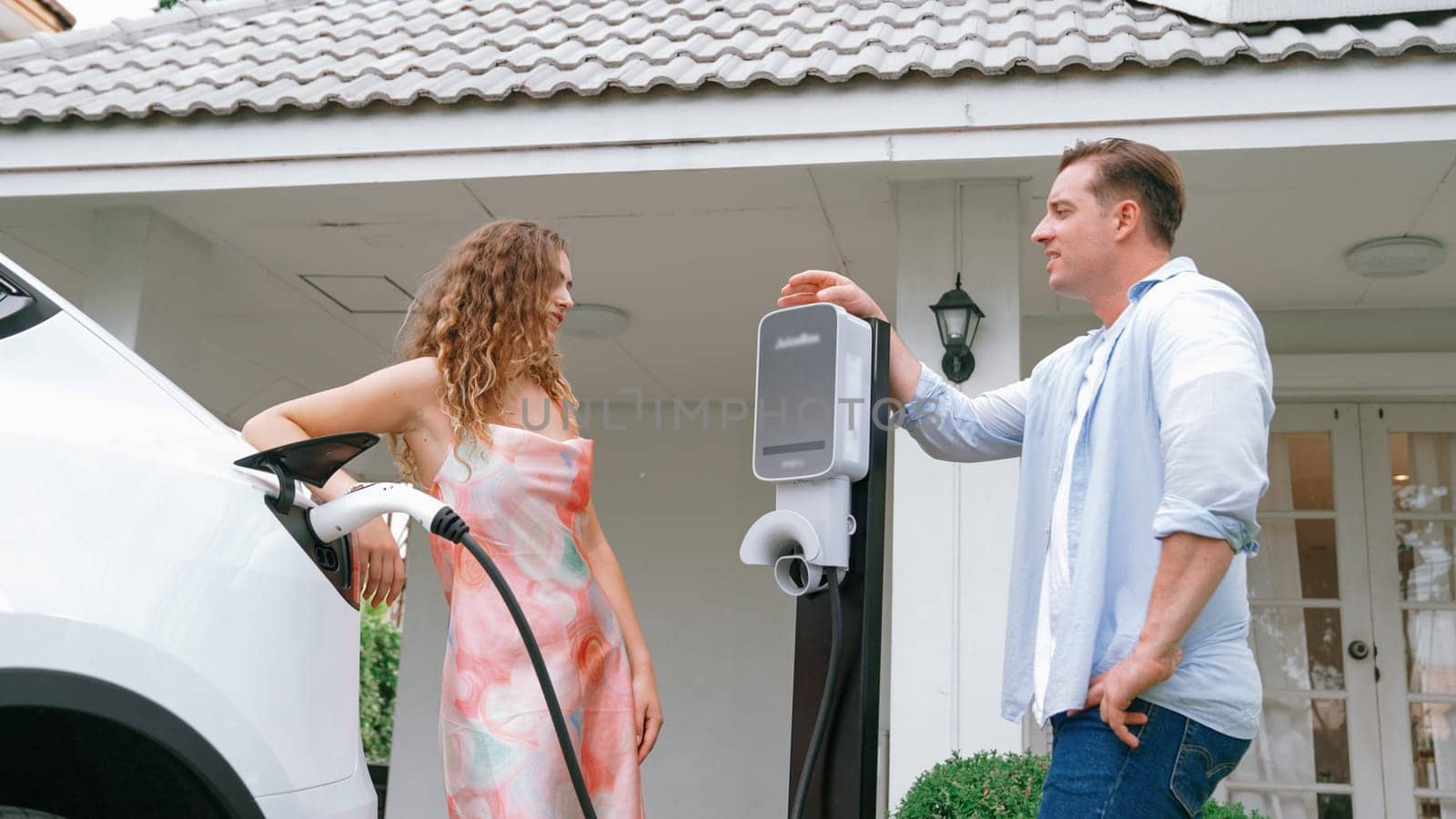 Happy and lovely couple with eco-friendly conscious recharging electric vehicle from EV charging station. EV car technology utilized as alternative transportation for future sustainability. Synchronos