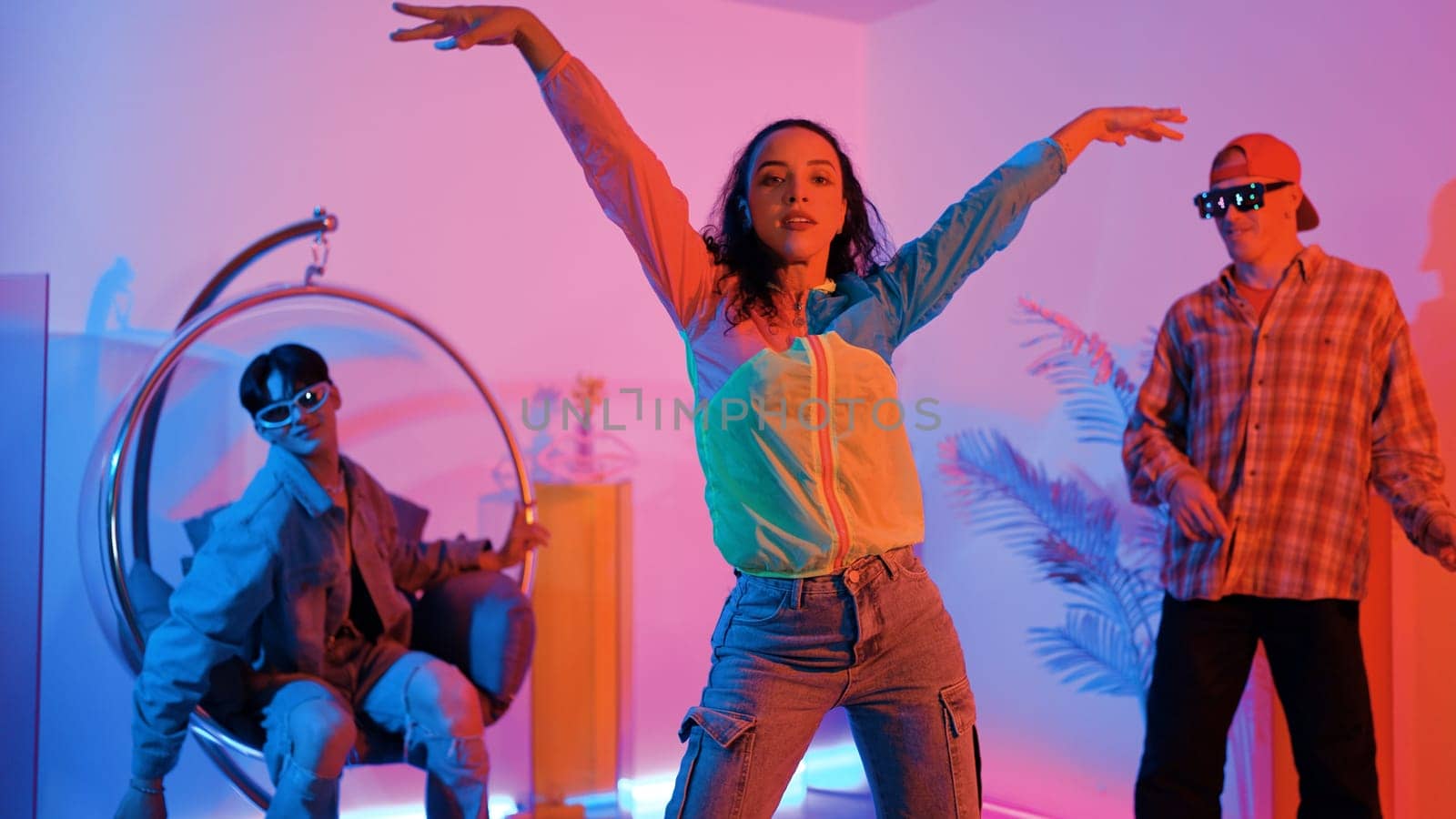 Attractive happy dancer looking at camera while smart woman moving at rhyme with neon light. Professional hispanic performer break dancing while wearing colorful cloth with diverse friend. Regalement.