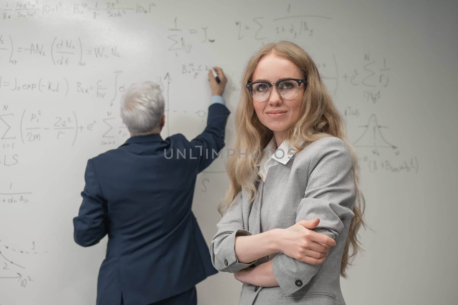 An elderly man writes on a white board and a young woman stands thoughtfully