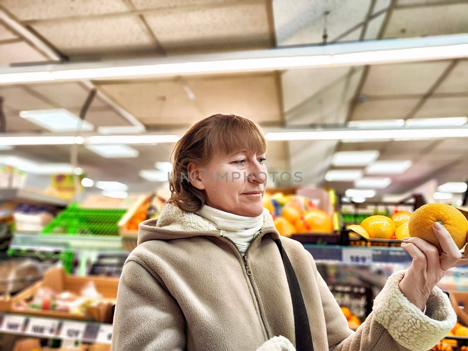 Middle-Aged Woman Holding an Orange in a Grocery Store. Woman examines an orange, considering nutrition in a store aisle. Concept of proper nutrition by keleny
