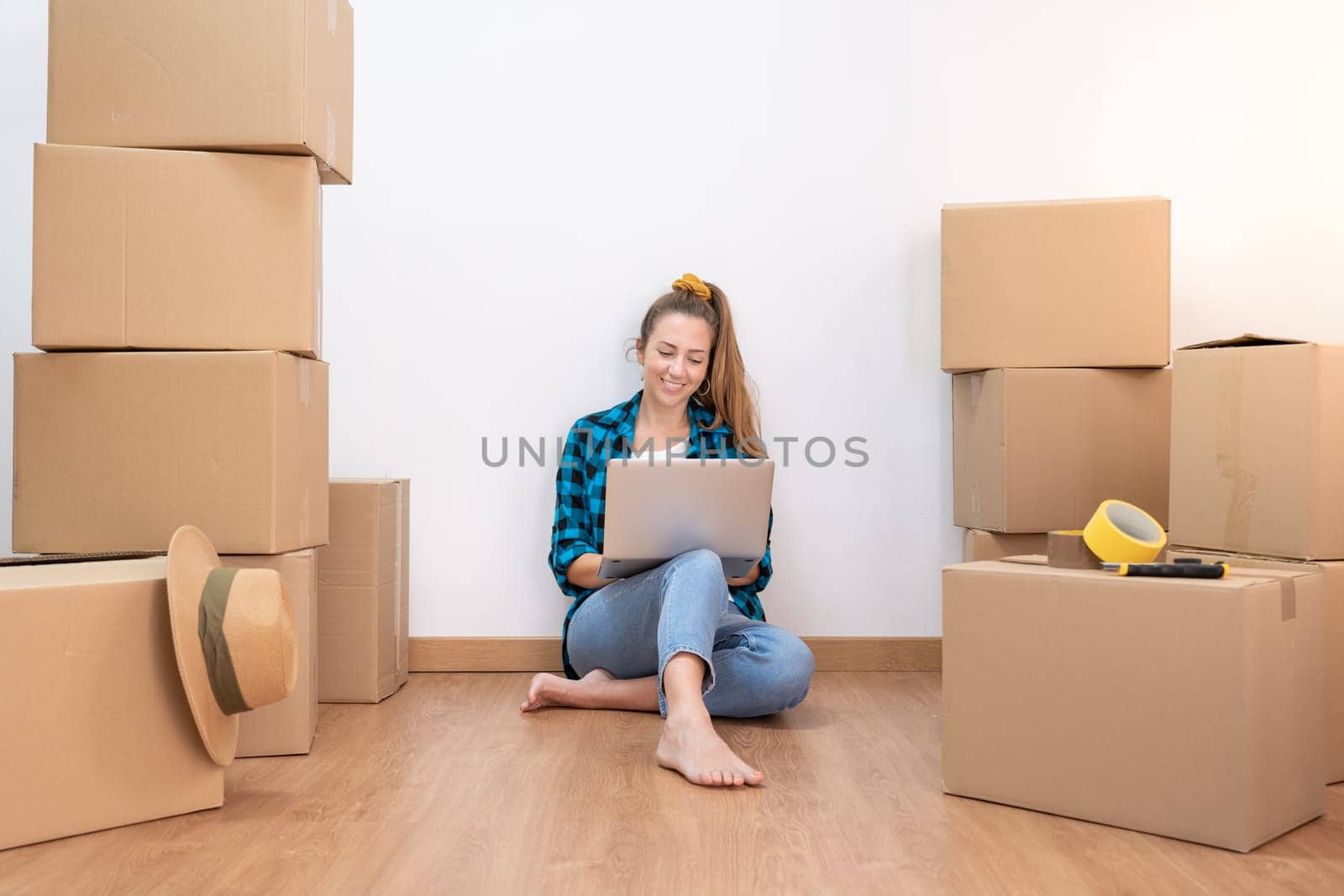 Young happy woman sitting on the floor with many boxes, working with computer in a new house. by PaulCarr