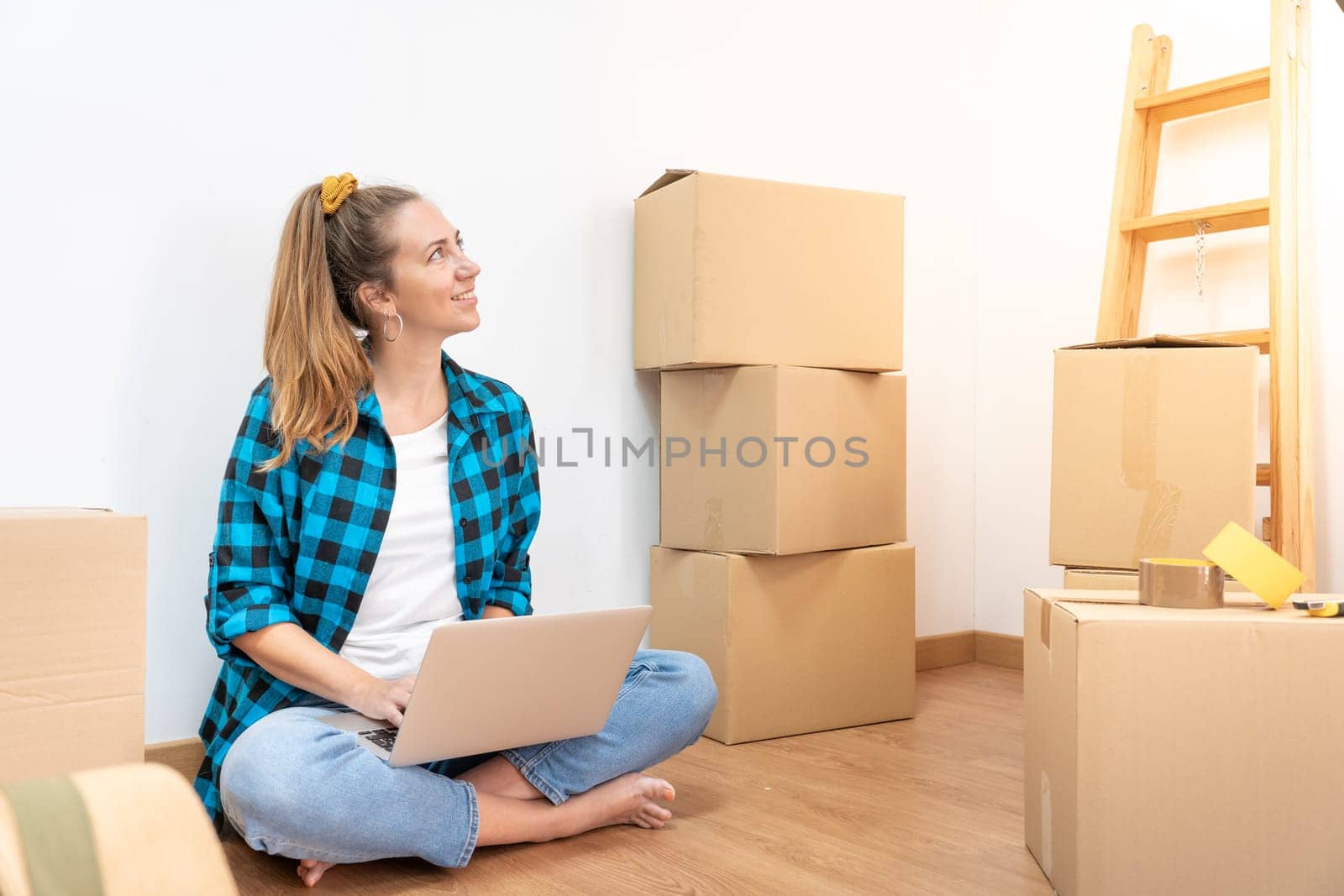 Young happy woman sitting on the floor with many boxes, working with computer in a new house. by PaulCarr