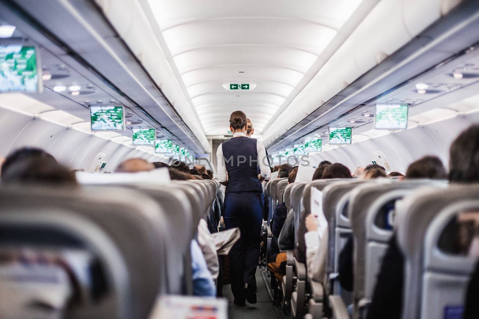 Interior of airplane with passengers on seats and stewardess in uniform walking the aisle, serving people. Commercial economy flight service concept