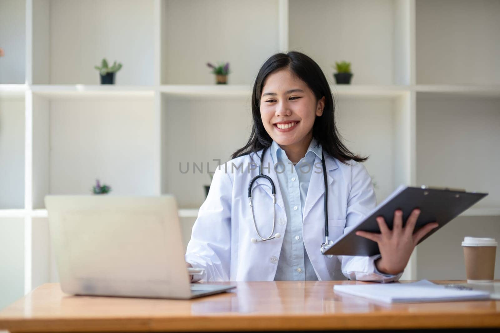 young asian lady doctor in white medical uniform with stethoscope using computer laptop talking video conference call with patient at desk in health clinic or hospital..