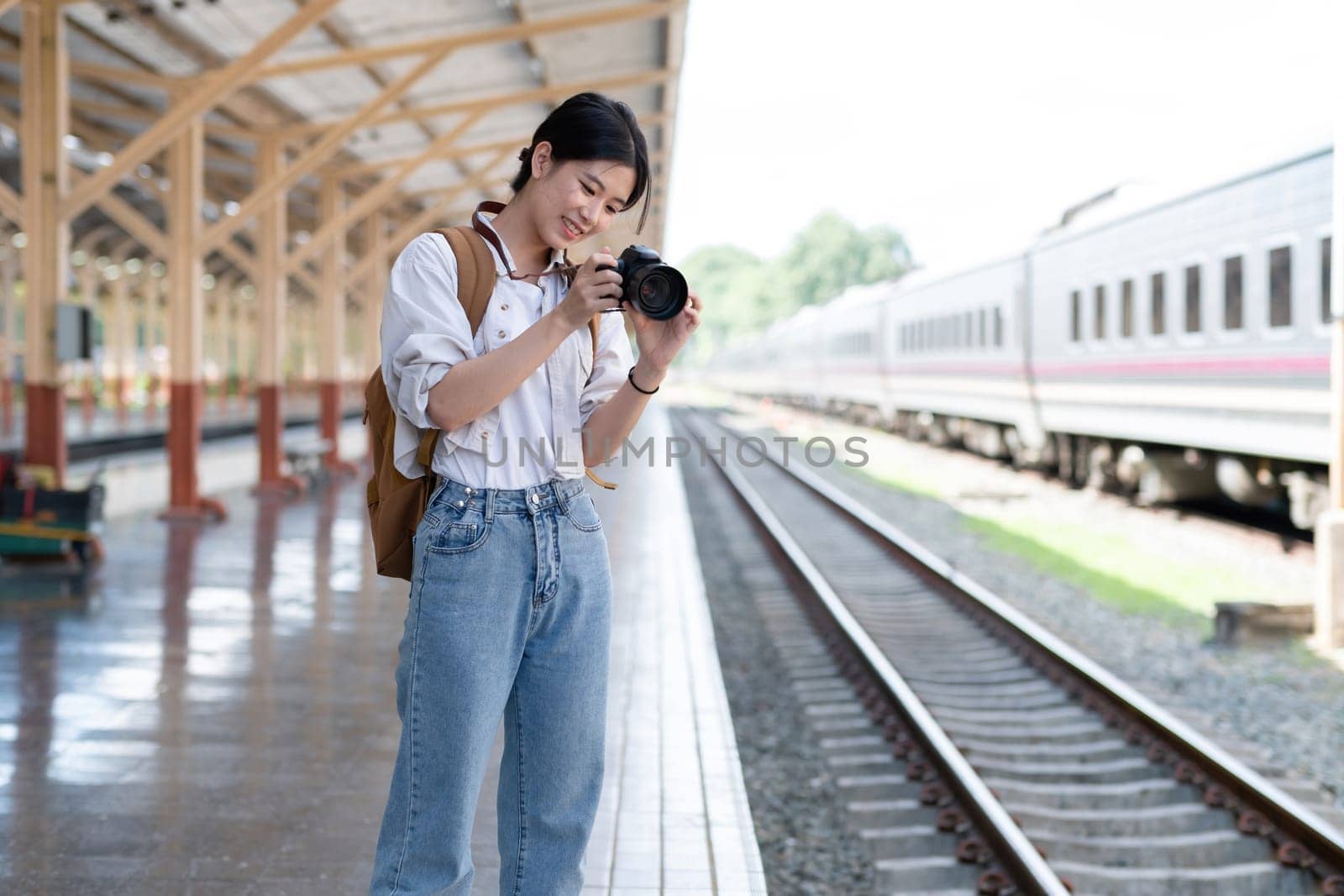 Young Asian woman holds a camera and takes photos of a train while waiting for the train at the train station to travel. by wichayada