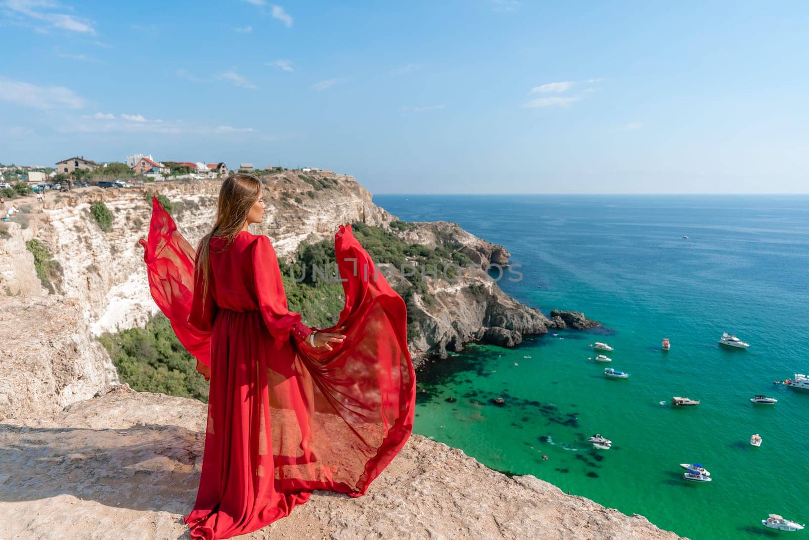 Red Dress Woman sea Cliff. A beautiful woman in a red dress and white swimsuit poses on a cliff overlooking the sea on a sunny day. Boats and yachts dot the background