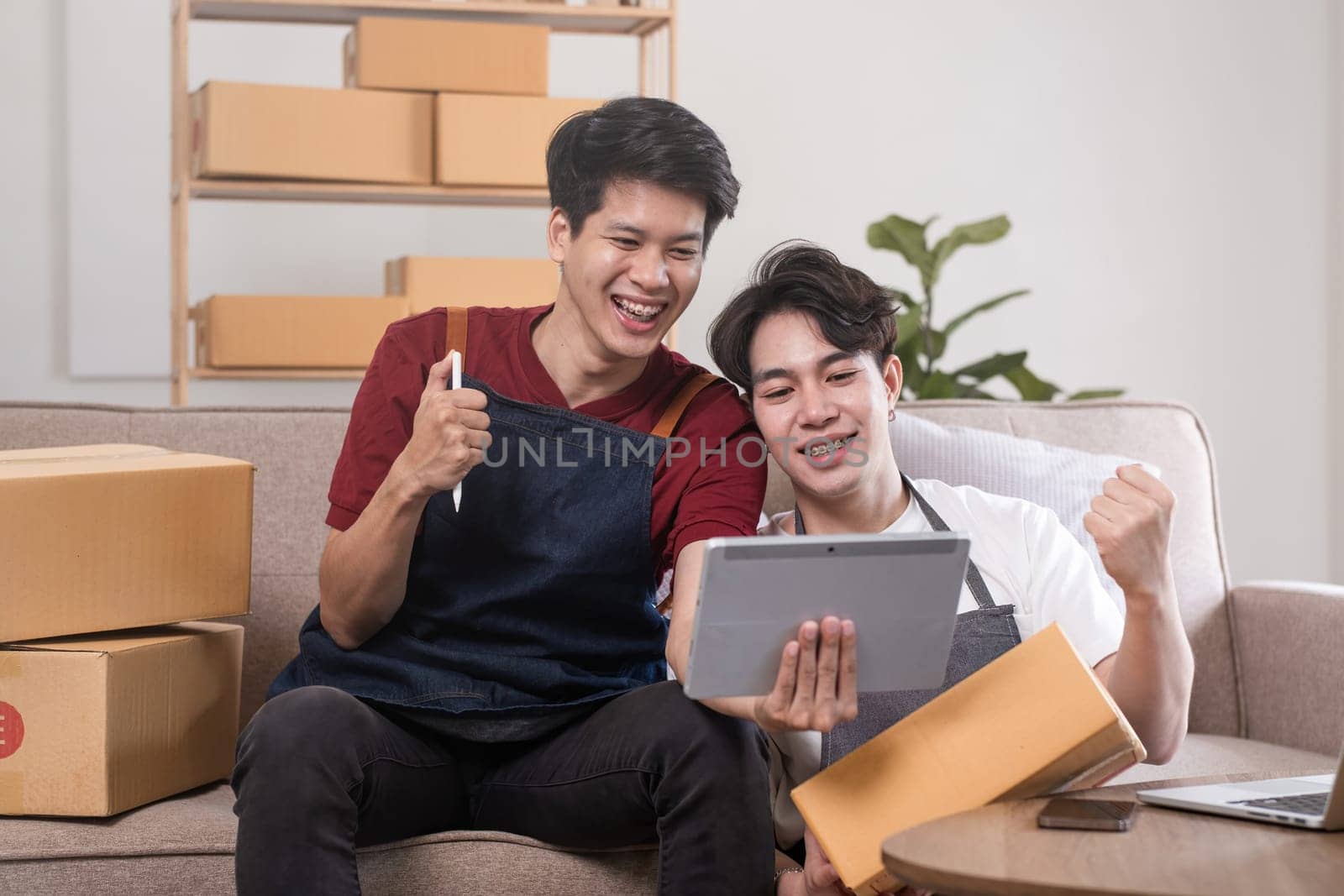A gay couple running a small business takes order on a laptop and sells product online together in a room full of boxes. by wichayada