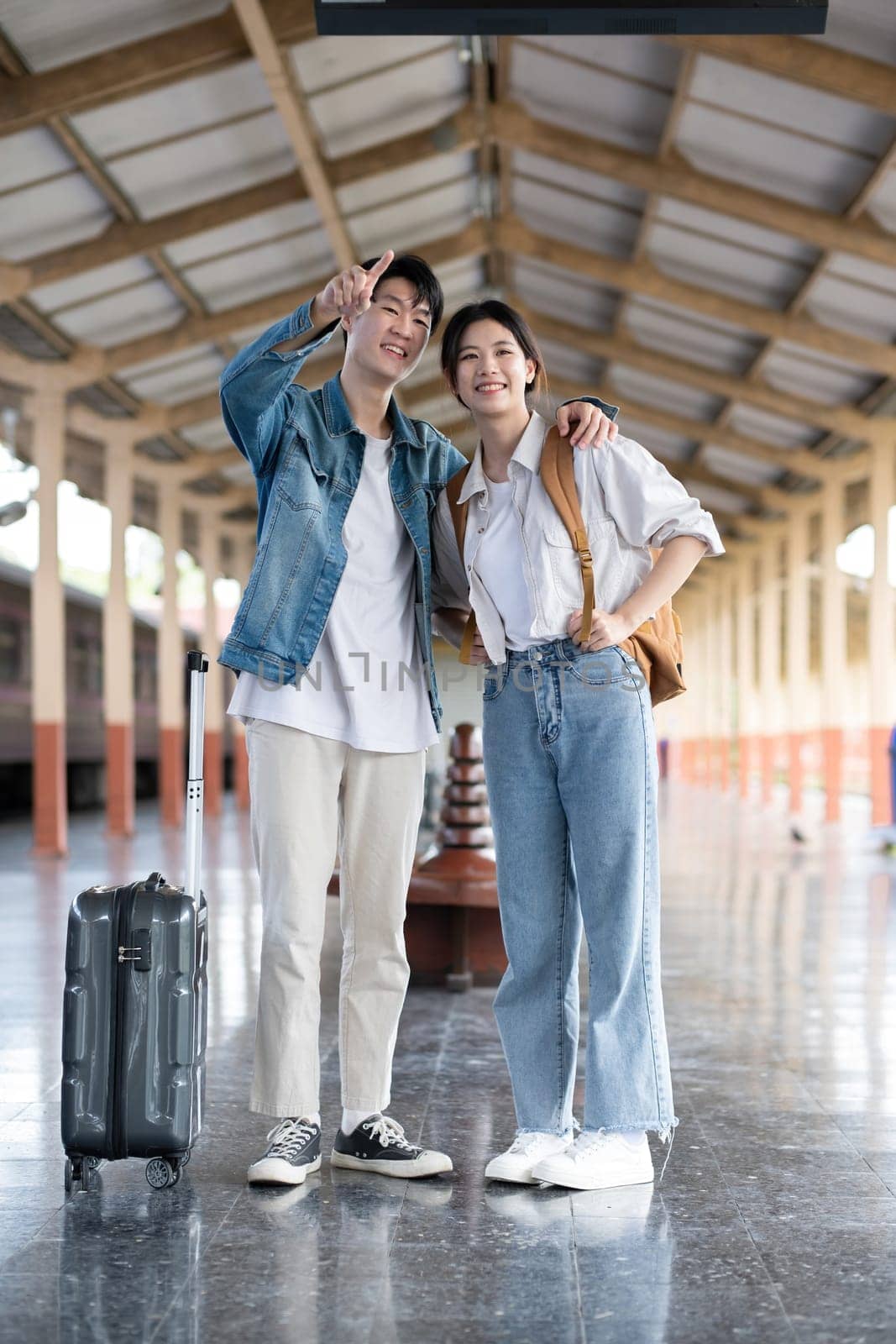 Happy Asian couple holding suitcases and camera preparing to wait for train at train station for vacation trip together..