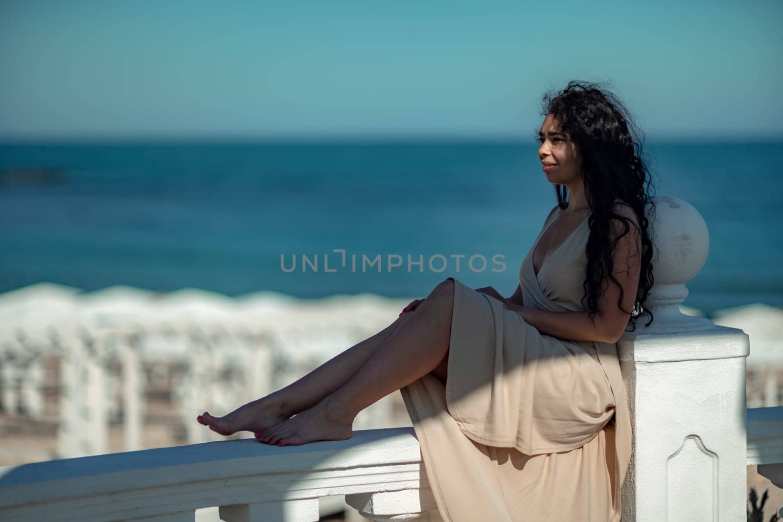 A woman in a tan dress is sitting on a white railing overlooking the ocean. The scene is serene and peaceful, with the woman enjoying the view and the calming sound of the waves. by Matiunina