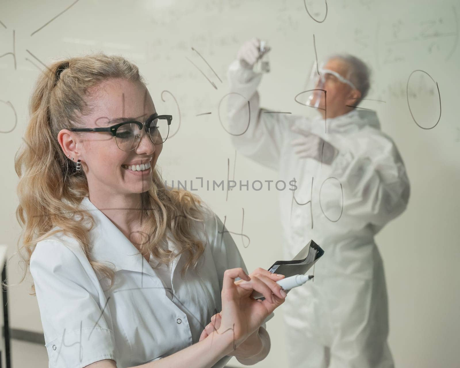A woman chemist writes a formula on glass. An elderly Caucasian man in a protective suit is doing tests