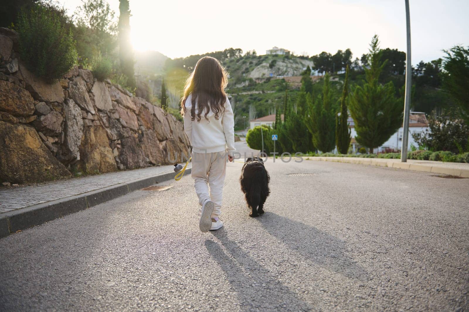 View from the back of a cute child girl in white sports clothes walking her dog on leash on the countryside against sunset sunbeams background. People, playing pets and nature concept