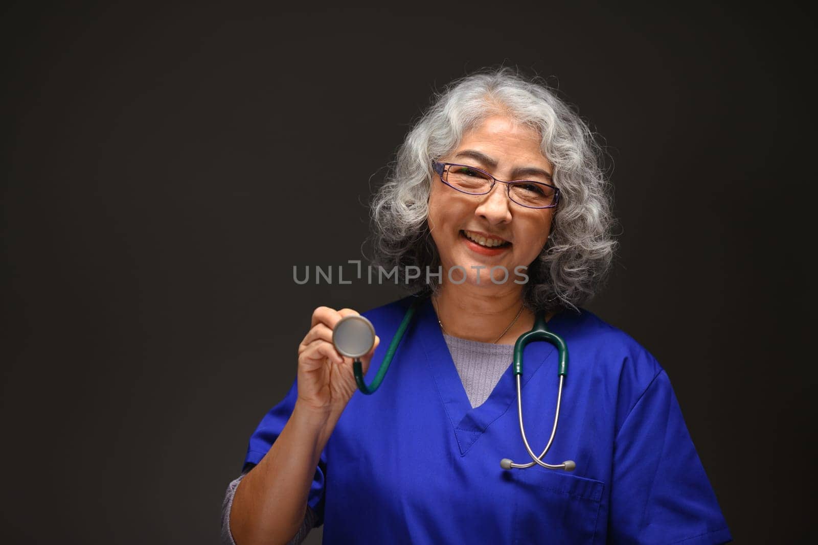 Portrait of friendly mature female doctor with stethoscope around her neck standing on black background.