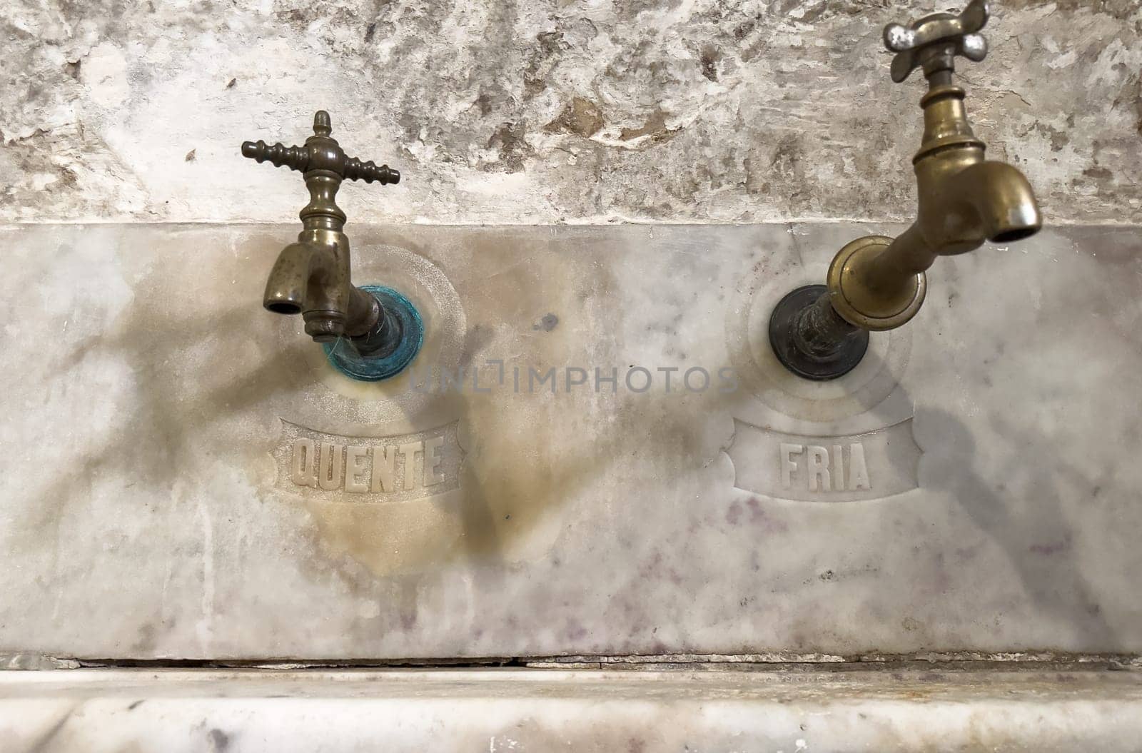 Vintage brass faucets marked QUENTE (hot) and FRIA (cold) on an old marble sink.