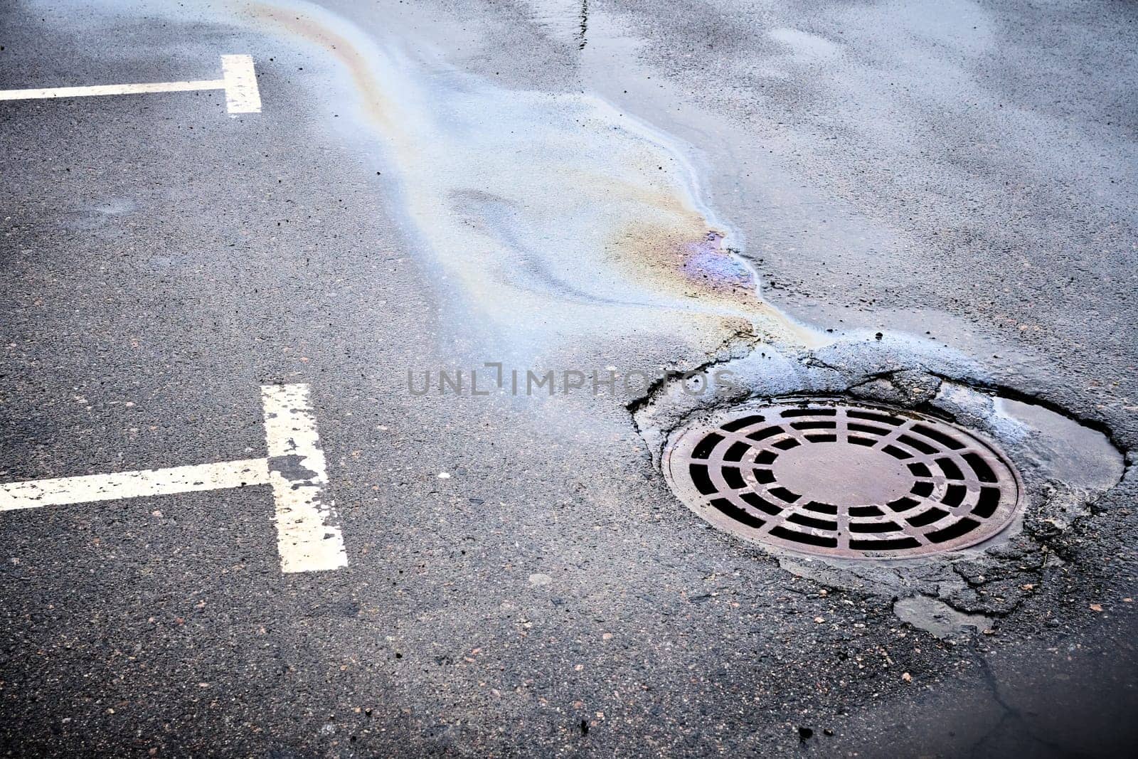 An oil slick of gasoline or oil on an asphalt road flows into a storm drain.