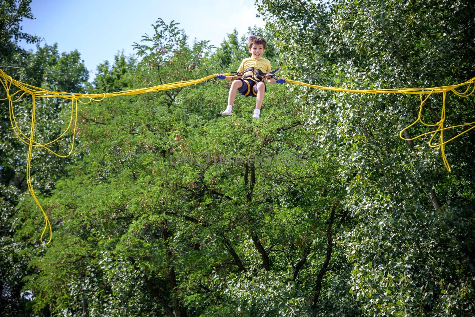 The boy is jumping on a bungee trampoline. A child with insurance and stretchable rubber bands hangs against the sky. The concept of happy childhood and games in the amusement park.