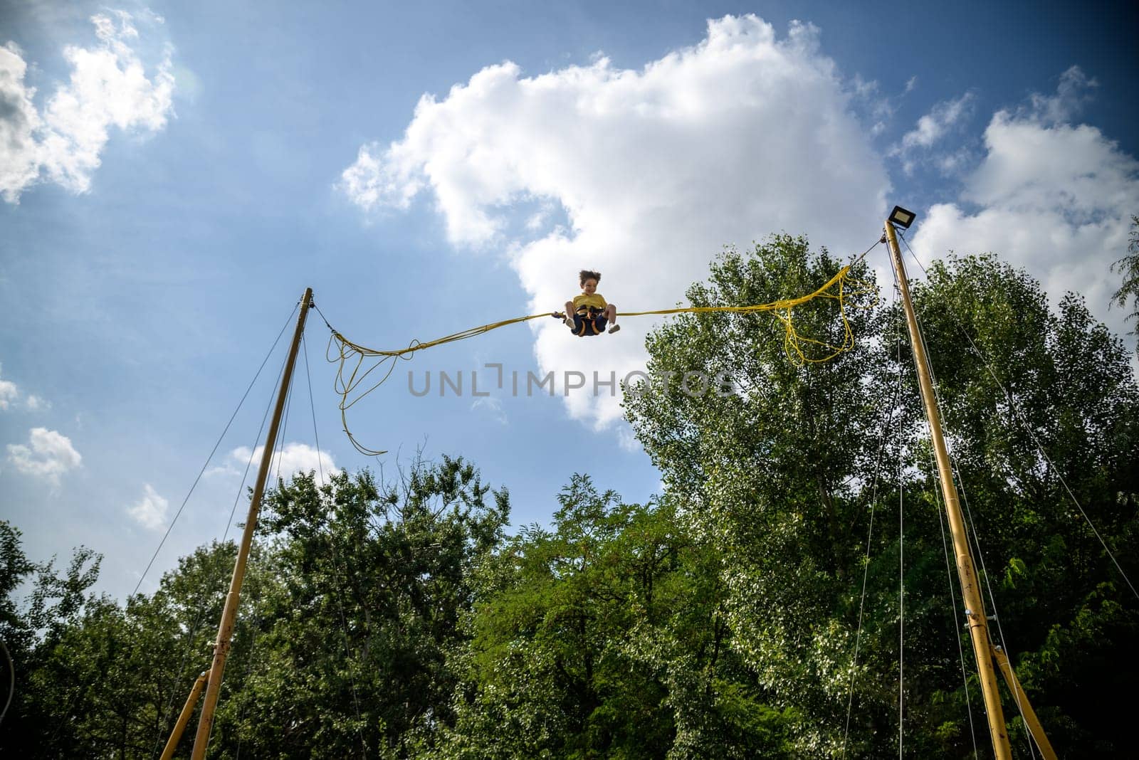 The boy is jumping on a bungee trampoline. A child with insurance and stretchable rubber bands hangs against the sky. The concept of happy childhood and games in the amusement park.