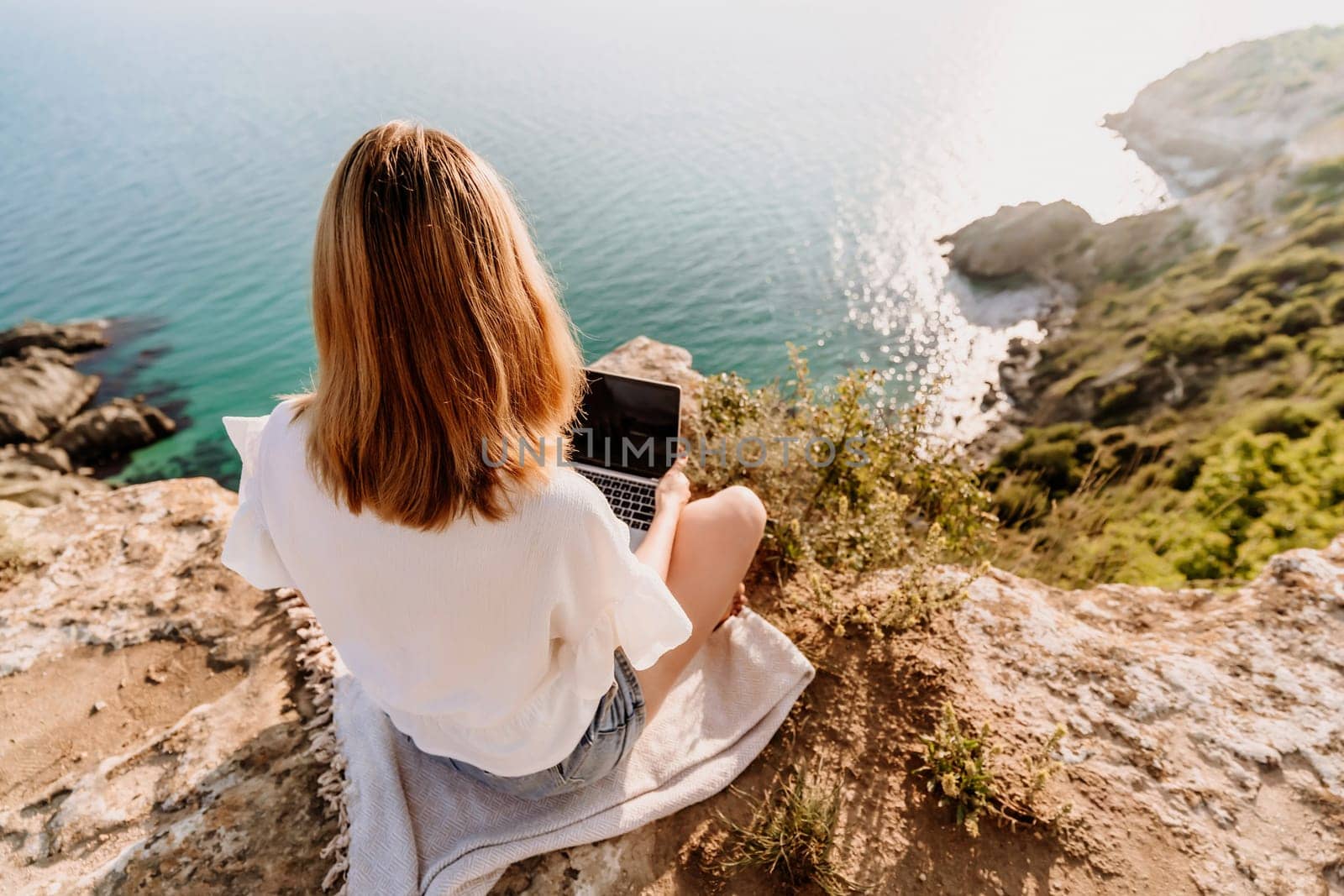 A woman is sitting on a rock overlooking the ocean with a laptop in front of her. She is enjoying the view and the peacefulness of the location