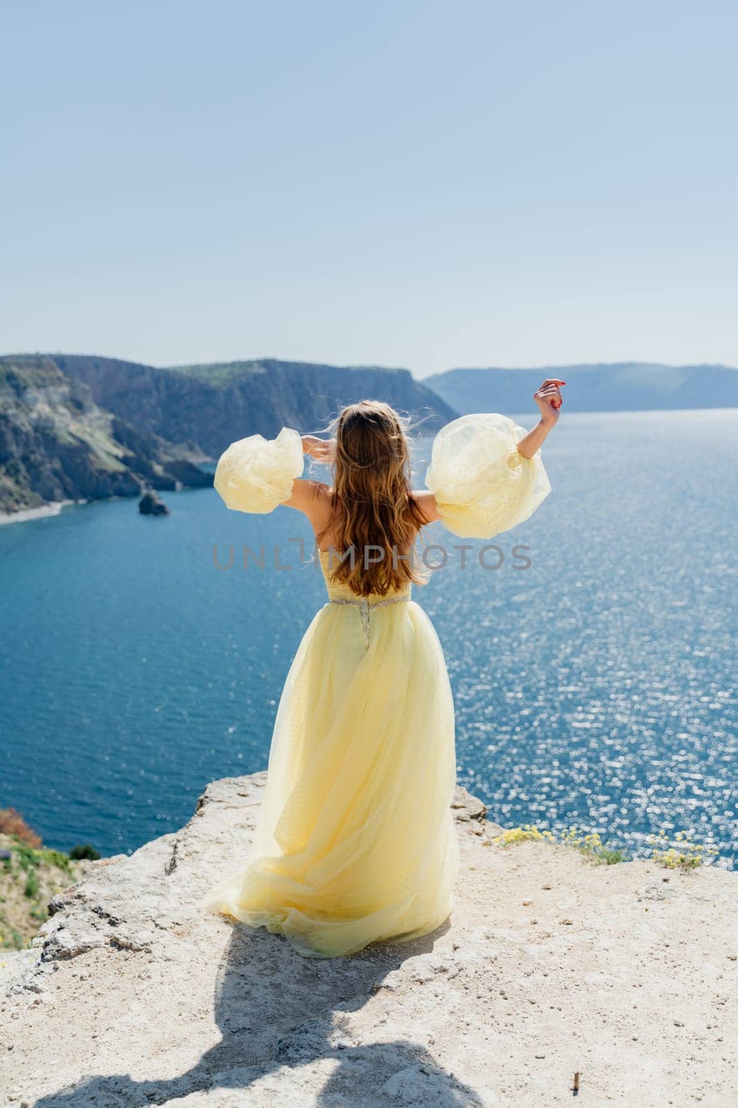 Woman in a yellow dress on the sea. Side view Young beautiful sensual woman in yellow long dress posing on a rock high above the sea at sunset. Girl in nature against the blue sky by Matiunina
