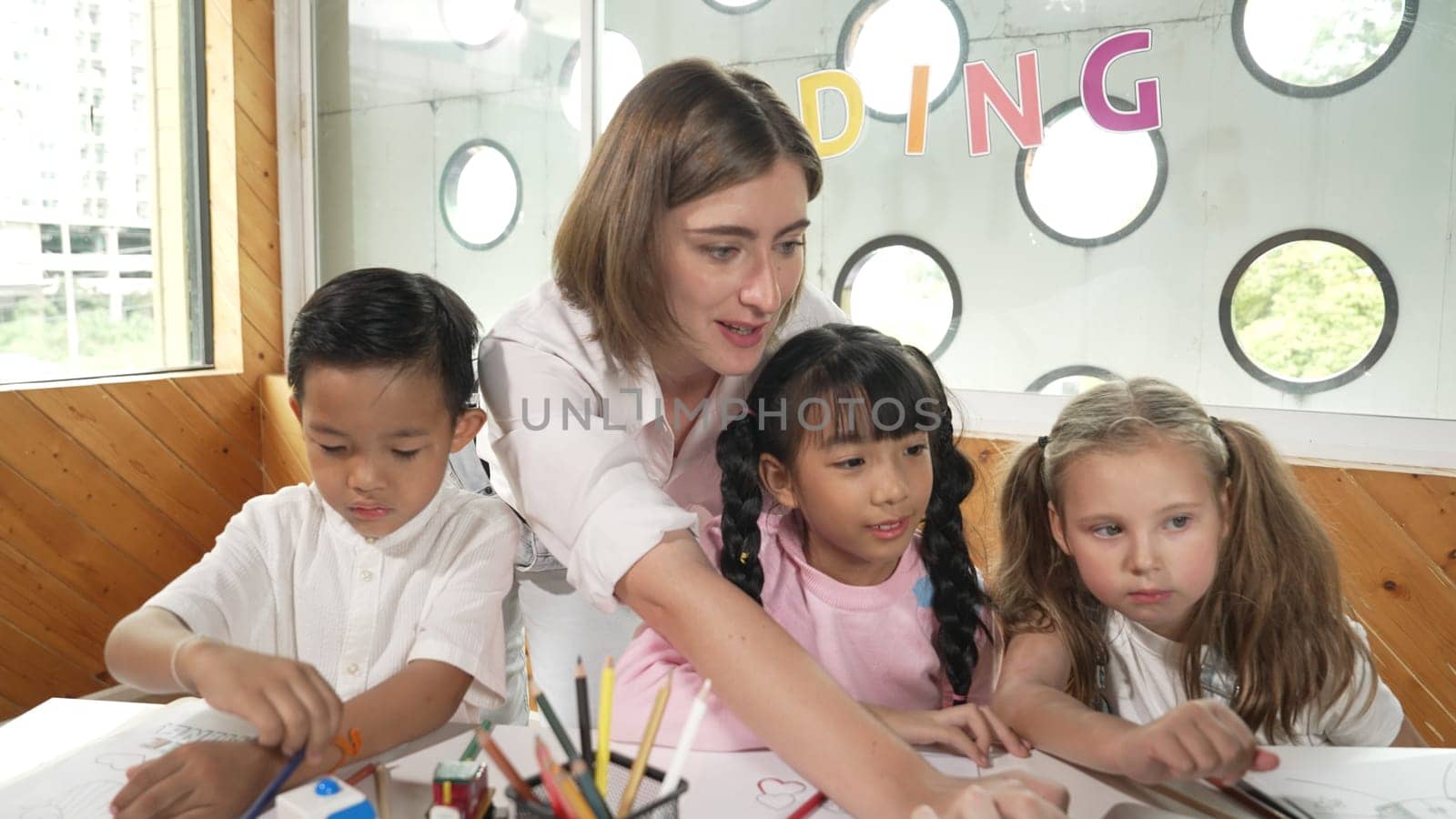 Teacher clicking at laptop while diverse student looking at screen. Children excited in presentation while drawing or writing art books at table with toys and color pencil placed on table. Erudition.