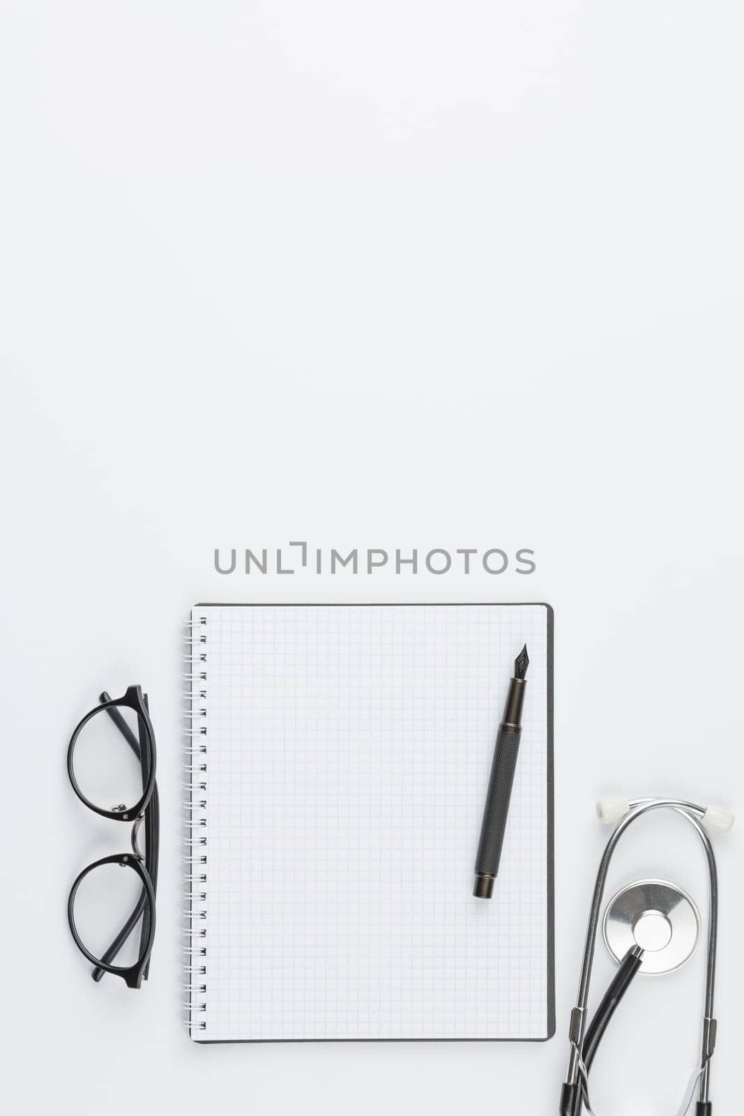 Stethoscope with glasses at workplace of medical worker. Doctor desktop top view. Spiral notebook with fountain pen isolated on white background. Doctor appointment concept, flat lay. Vertical photo.