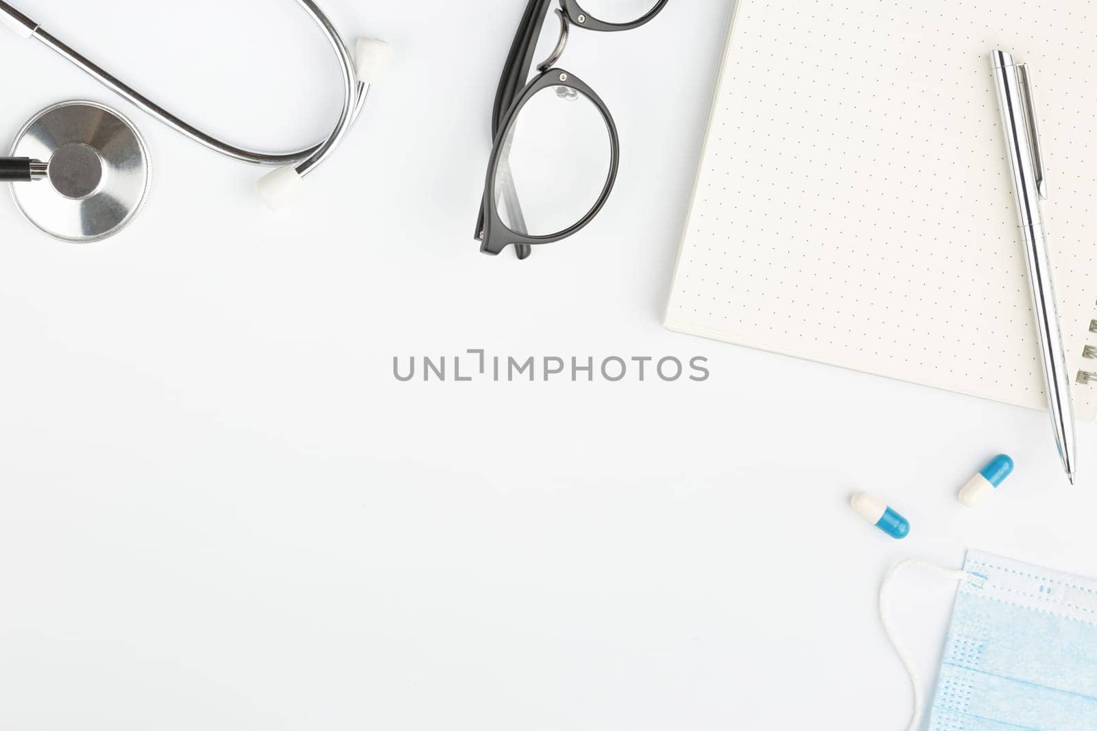 Open spiral notebook with pen and glasses flat lay. Stethoscope with medical mask and pills on white isolated background top view. Doctor's desktop concept.