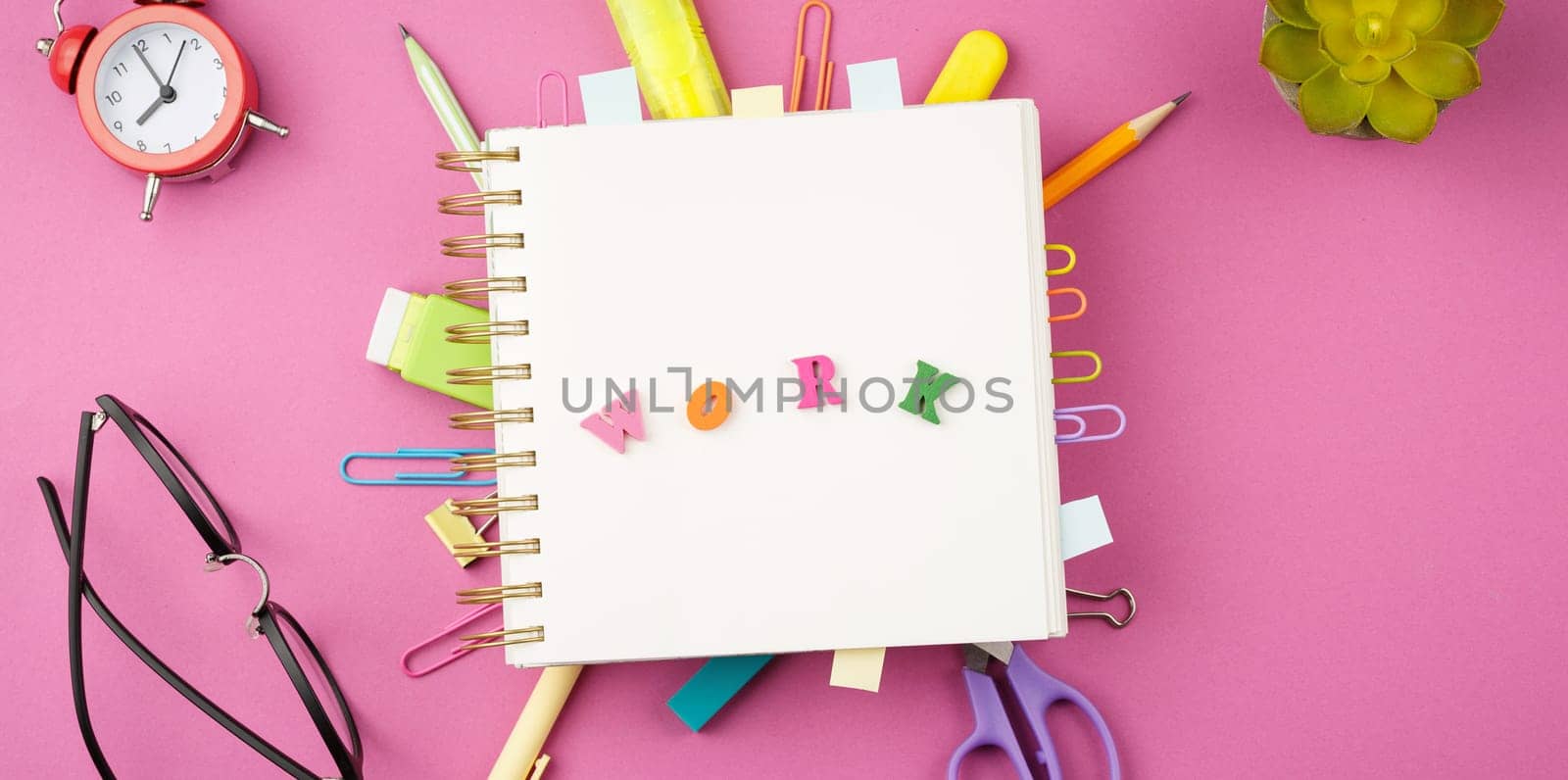 Open spiral notebook with bookmarks from paper clips and leaves for notes, pencil, scissors, binders, alarm clock, glasses on a pink background. Top view. Stationery concept. Office tools.