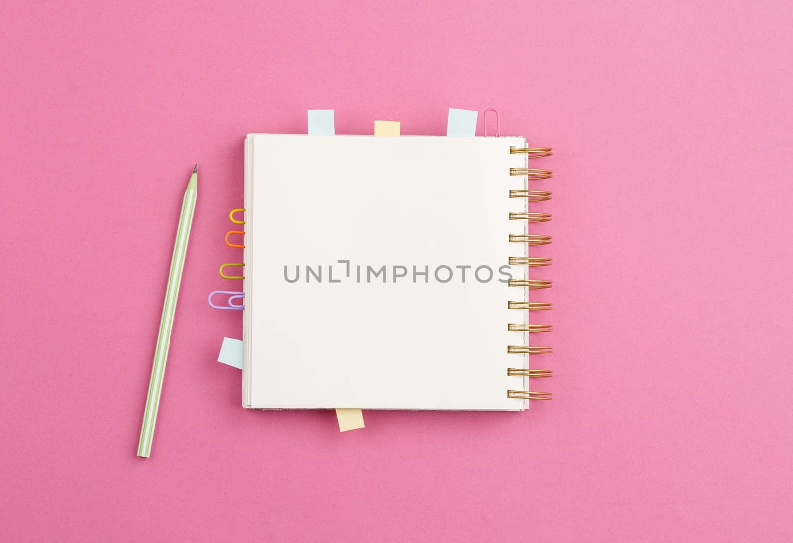 Open spiral notebook with bookmarks from paper clips and leaves for notes, pencil on a pink background. Top view.