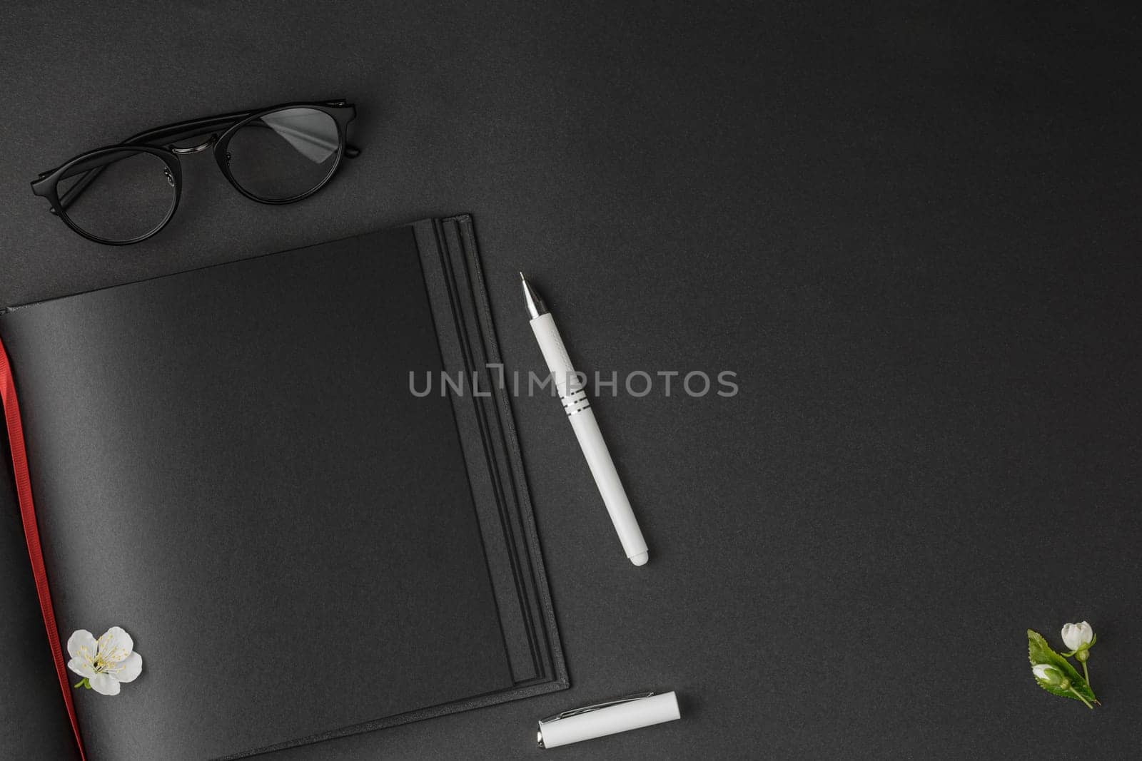 Open notebook with red bookmark and black sheets, white pen, glasses and white flowers on dark isolated background flat lay.