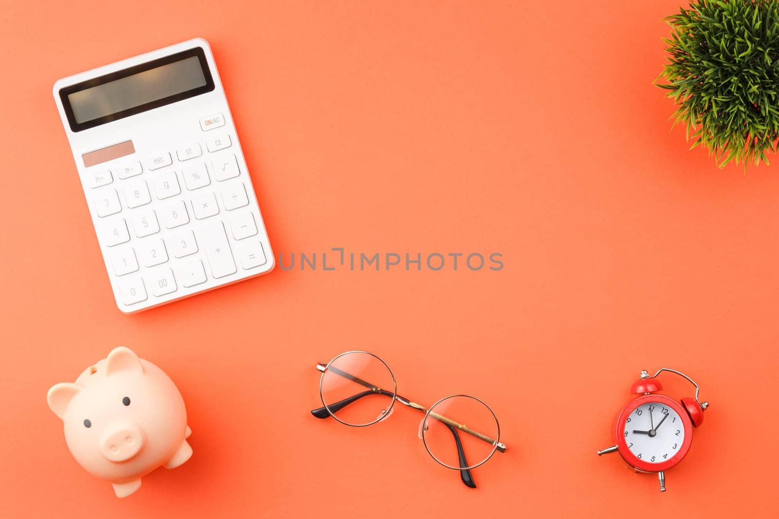 White calculator and pig piggy bank, alarm clock, glasses and indoor plant on orange isolated background flat lay top view.