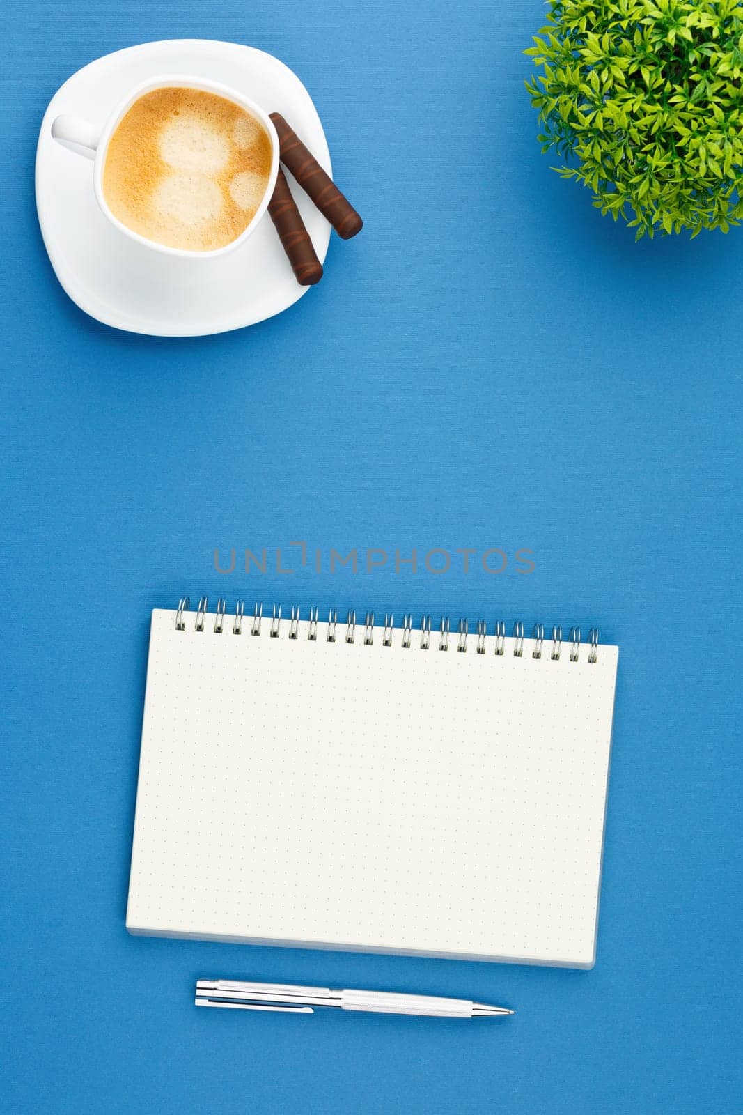 Spiral open notebook with a metal pen, dessert chocolate sweets, cup of coffee and home plant with green leaves on blue background. Flat lay, top view, Vertical photo.