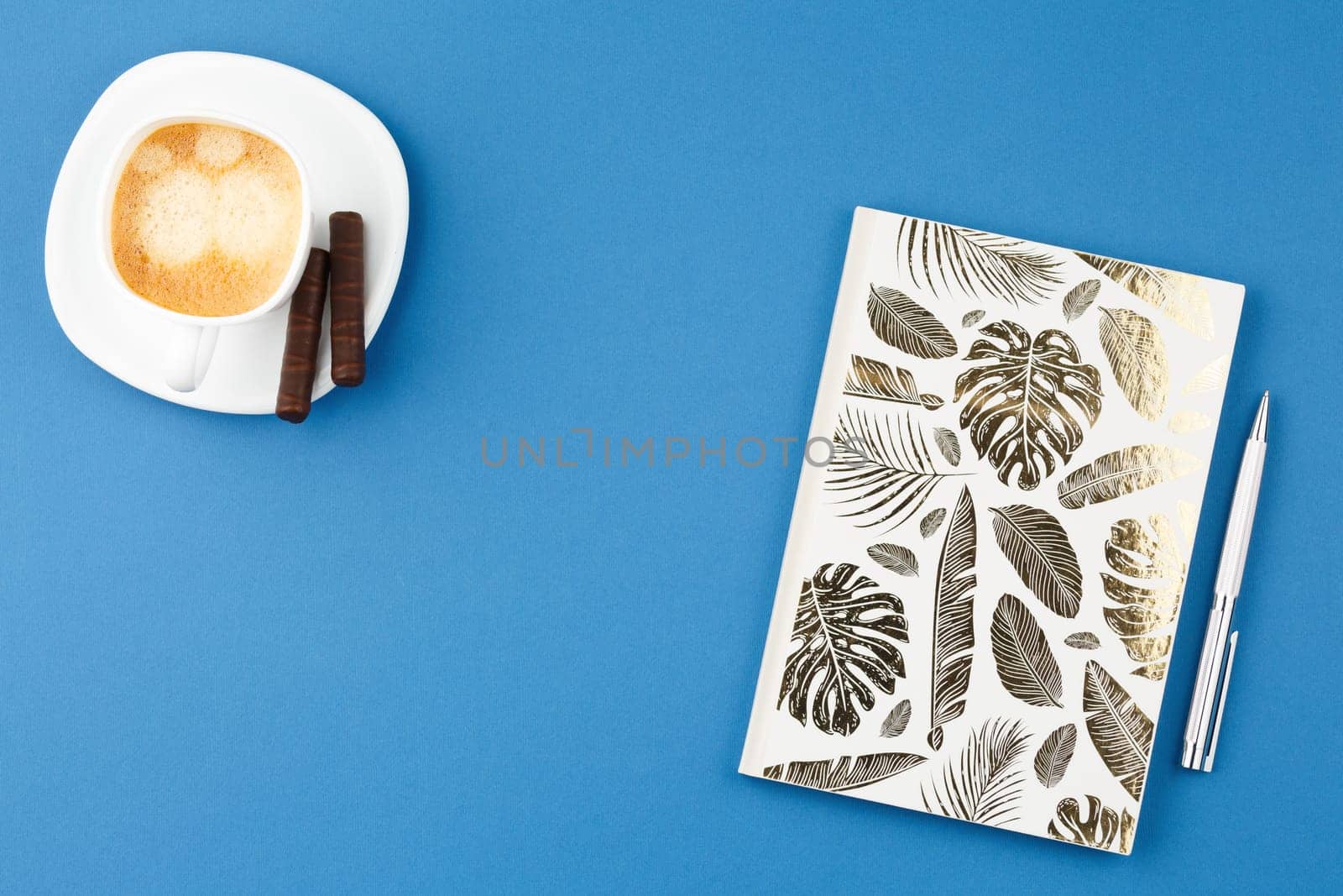 Notebook with metal pen, dessert chocolate sweets and cup of coffee on blue background. Flat lay, top view.
