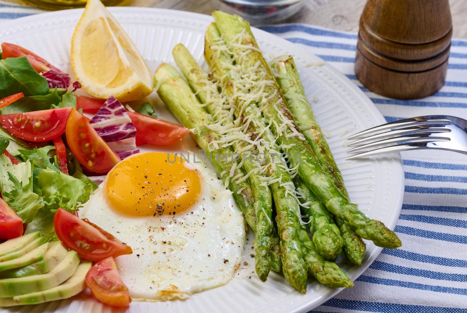 Round plate with cooked asparagus, fried egg, avocado and fresh vegetable salad on the table by ndanko