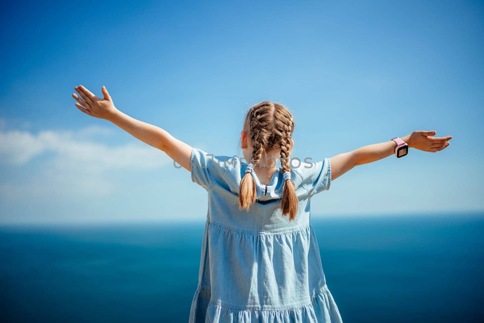 A young girl with pigtails is standing on a beach, looking out at the ocean. She is wearing a blue dress and has her arms outstretched, as if she is embracing the beauty of the ocean