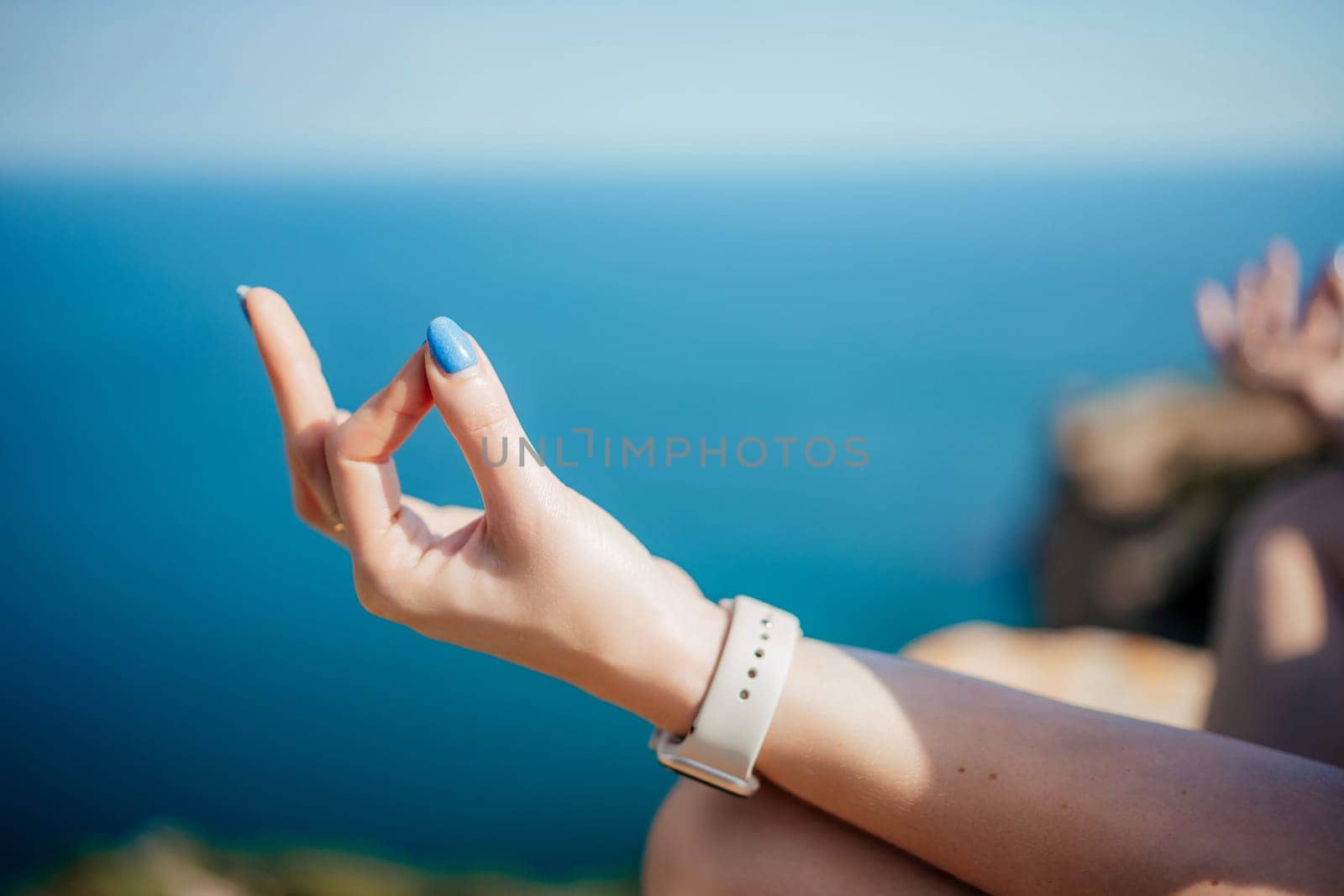 A woman is sitting on a rock by the ocean, with her hand in a prayer position. She is wearing a white watch and has blue nail polish on her nails. Concept of relaxation and mindfulness
