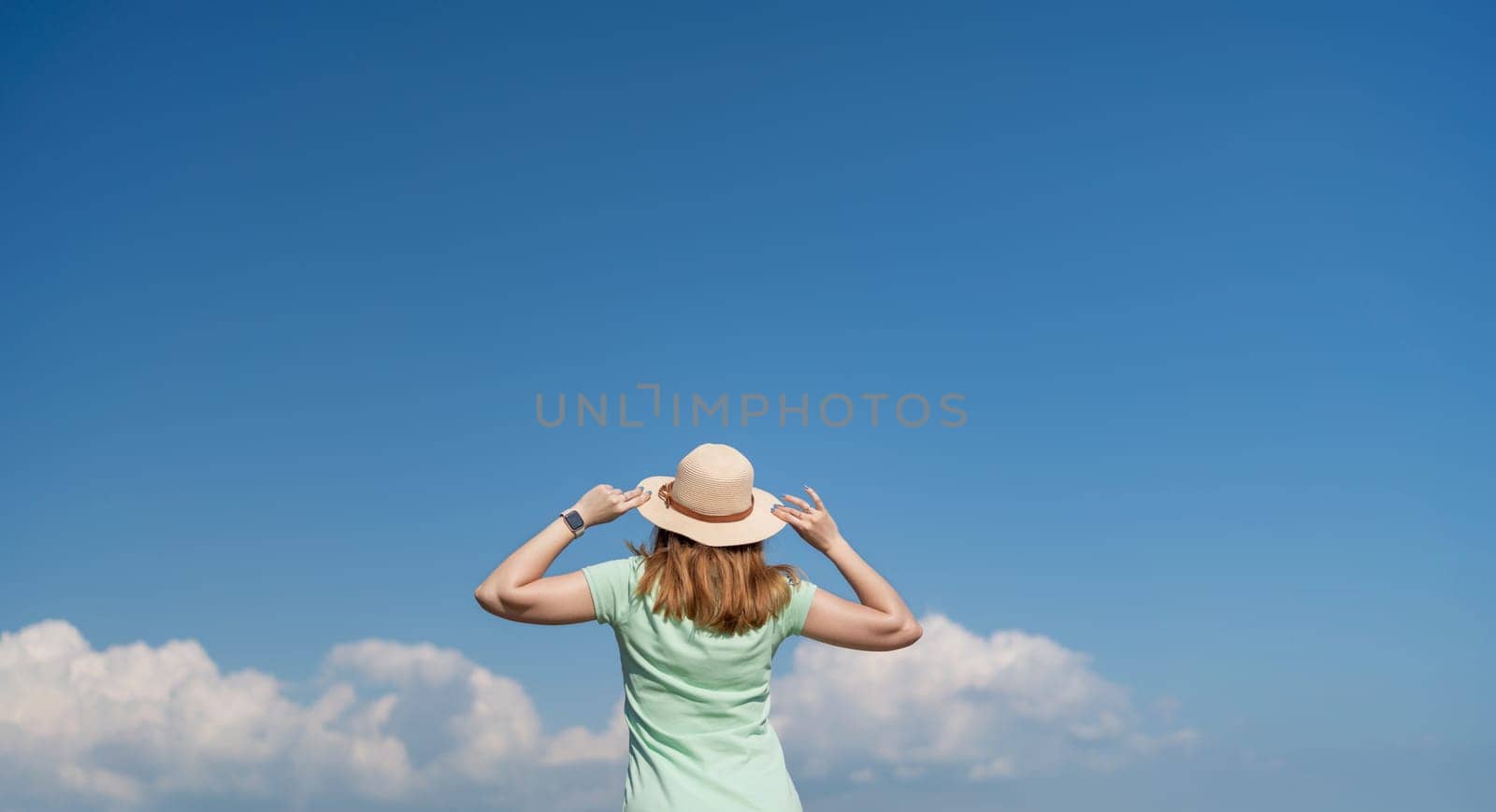 A woman wearing a straw hat is standing in a field with a clear blue sky above her. She is looking up at the sky, possibly admiring the clouds or just enjoying the view