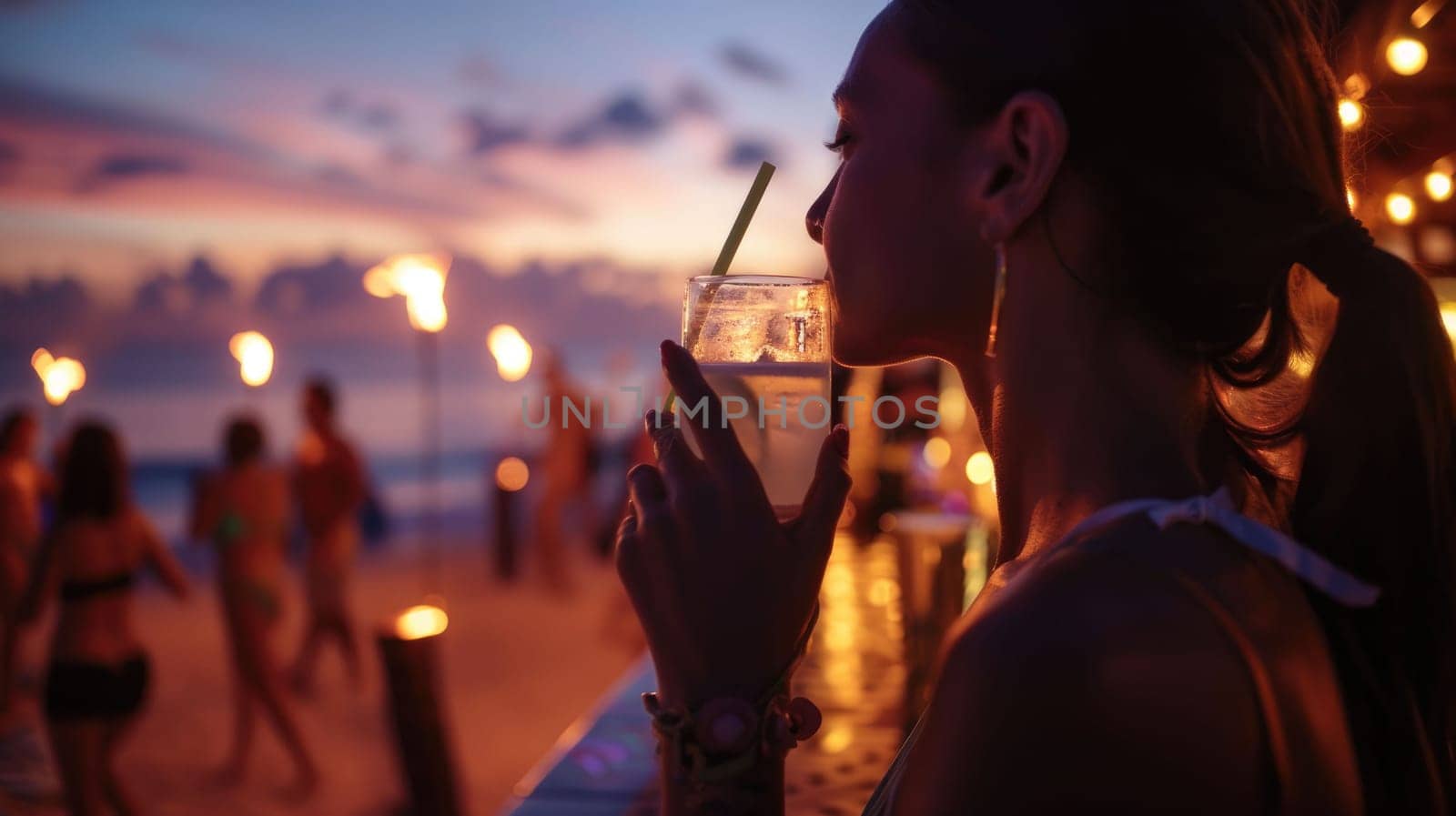 Under the midnight sky, a woman is enjoying a refreshing drink of water through a straw on the beach. The sound of music adds to the fun atmosphere during this latenight beach event AIG50