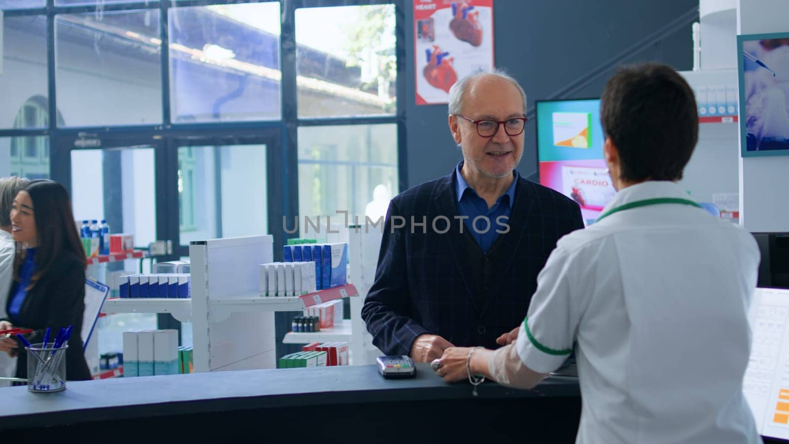 Senior shopper arriving at drugstore checkout counter after finding needed pharmaceutical items, purchasing pain relieving pills with credit card, being helped by chemist assistant with shopping bag